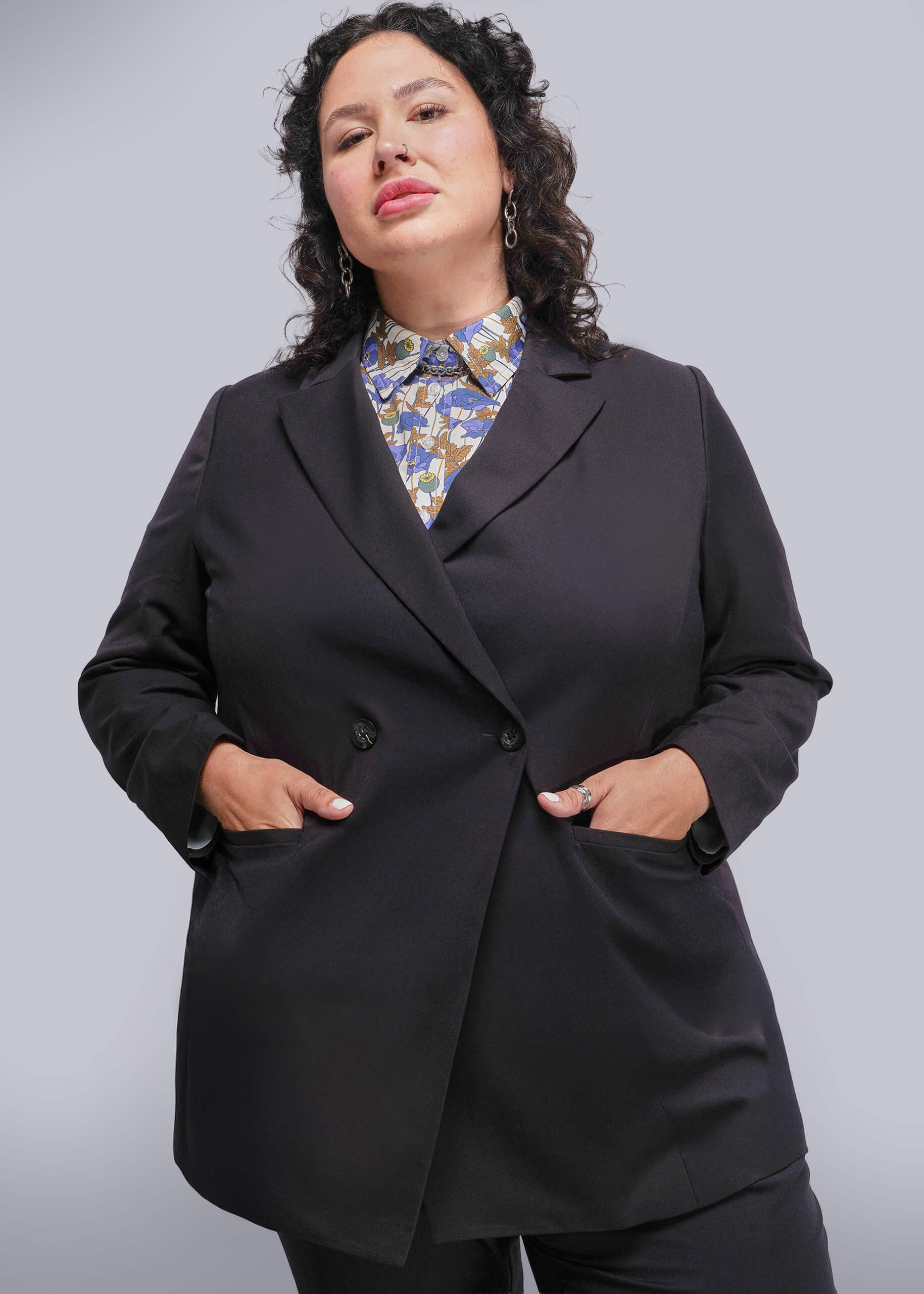 1X=Person wearing a black blazer over a patterned shirt on a plain background