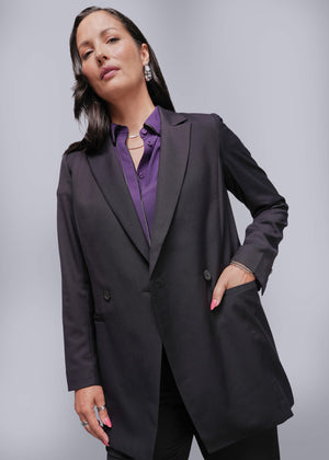 S=Woman wearing a black blazer over a purple shirt against a gray background