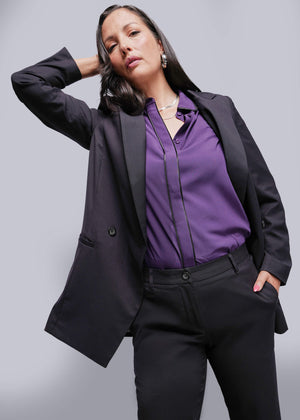 S=Woman wearing a black blazer and purple shirt against a gray background