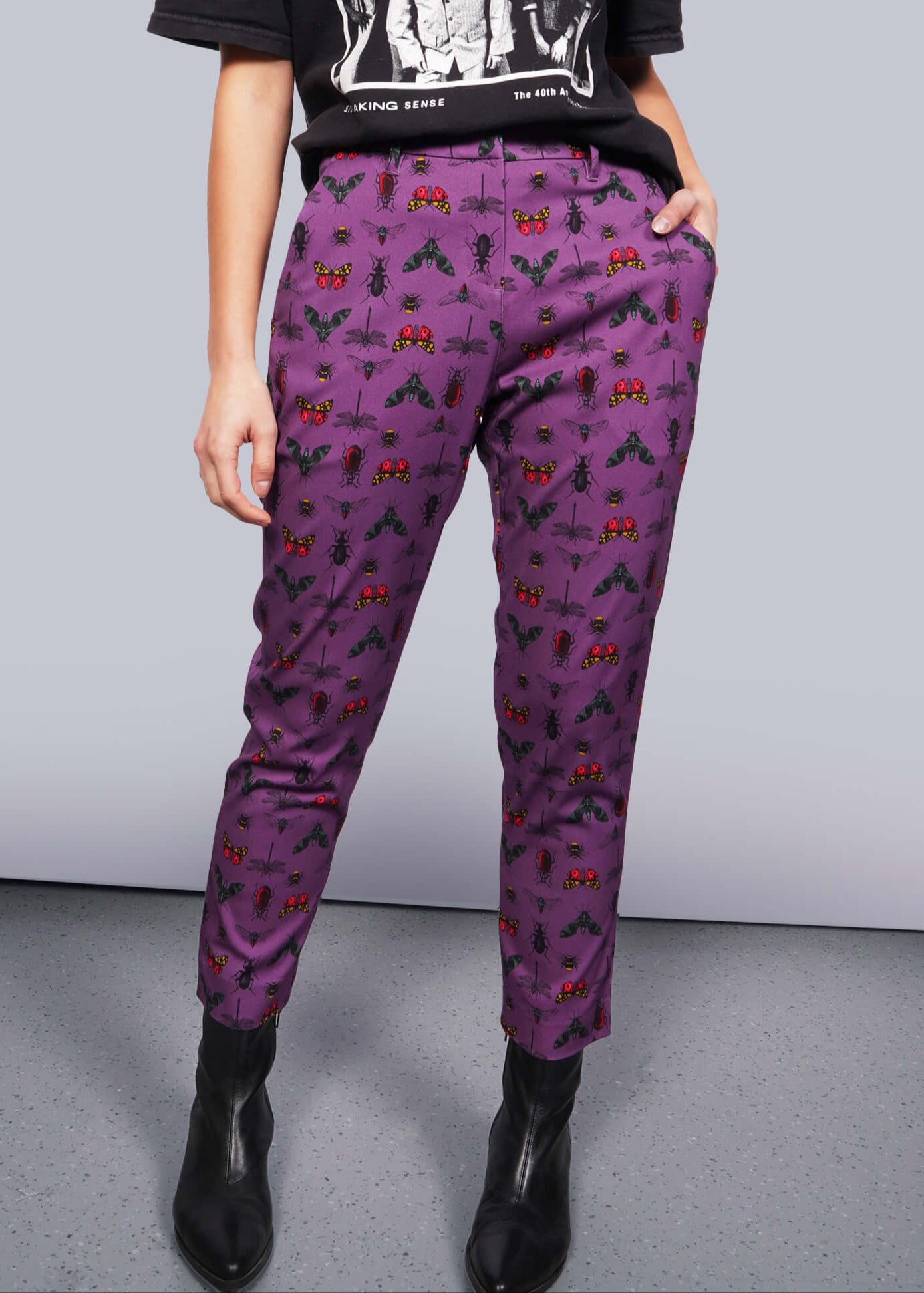 S= Pictured from the waist down, model stands in front of white studio backdrop wearing black graphic tee, black boots, and The Empower Slim Crop Pant in Insect Grape, a purple fabric with colorful insects printed on it.