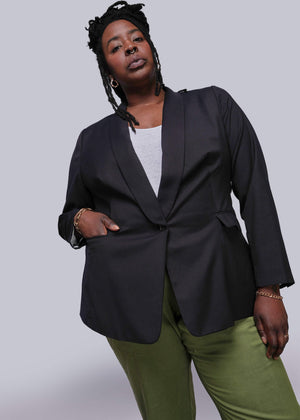 1X=Person wearing a black blazer and green pants against a gray background