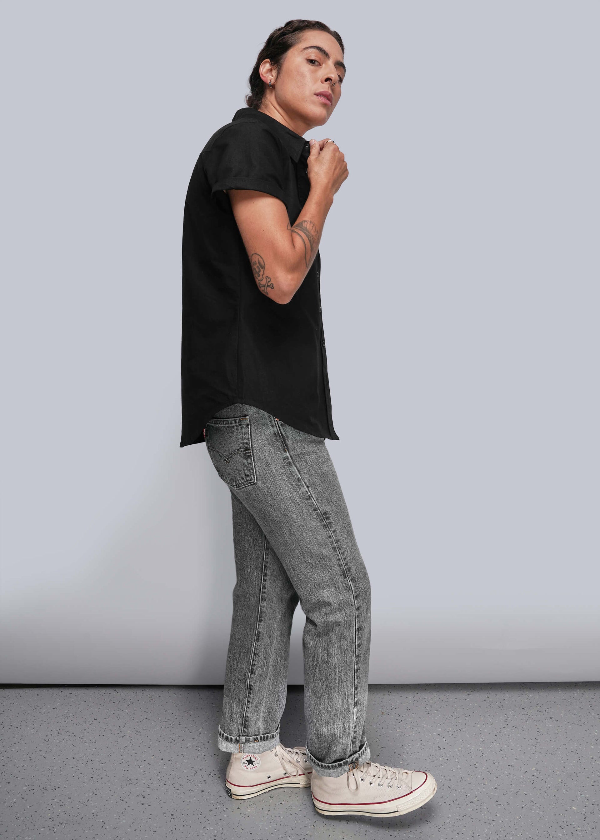 S=Person wearing a black shirt, gray jeans, and white sneakers against a plain background