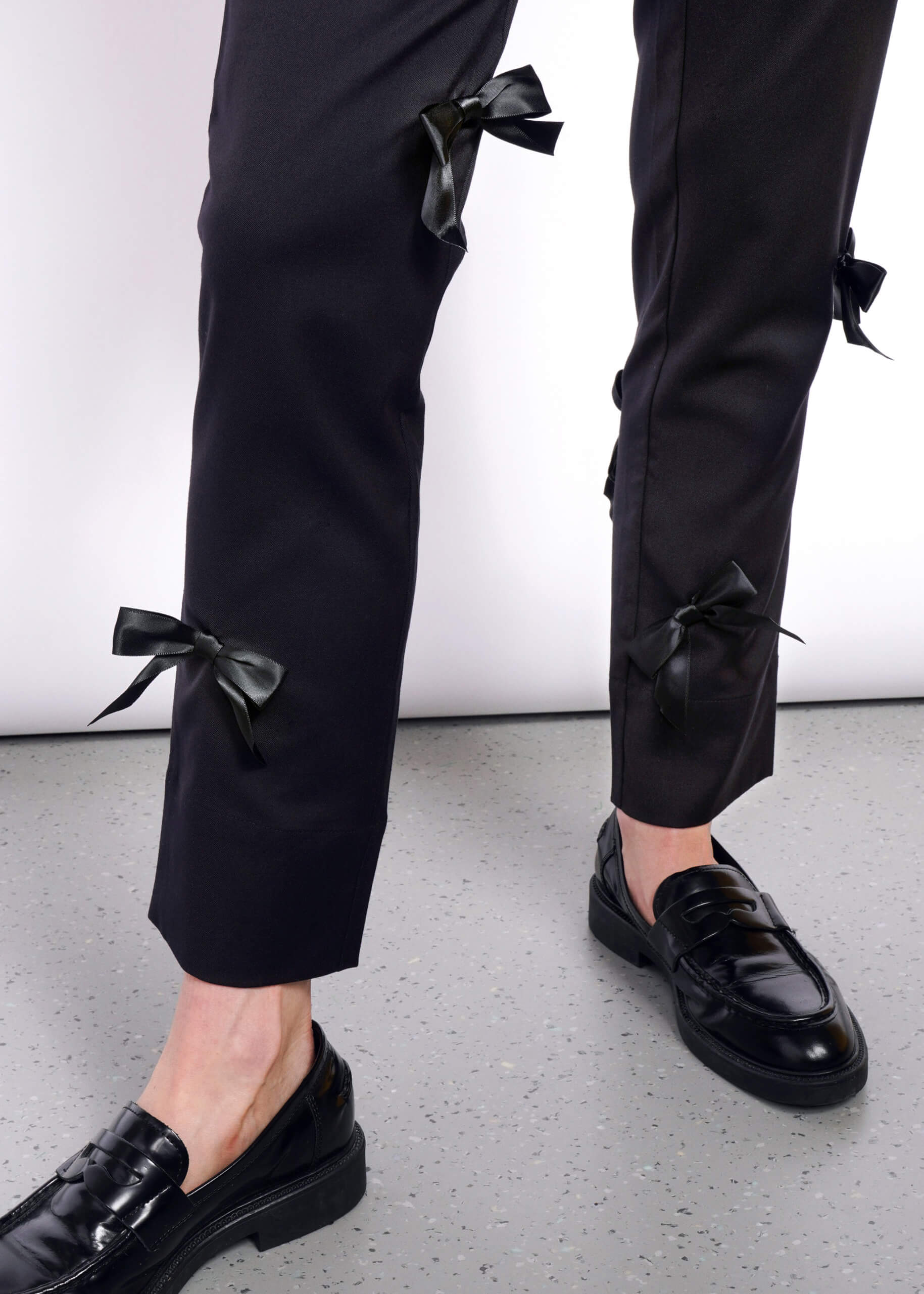 S=Someone wearing The Empower Bows Trouser, featuring mid-rise, black pants with exquisite bow embellishments on the sides, paired with shiny black loafers. The simple white background and speckled gray floor enhance the ultra-lux feel.