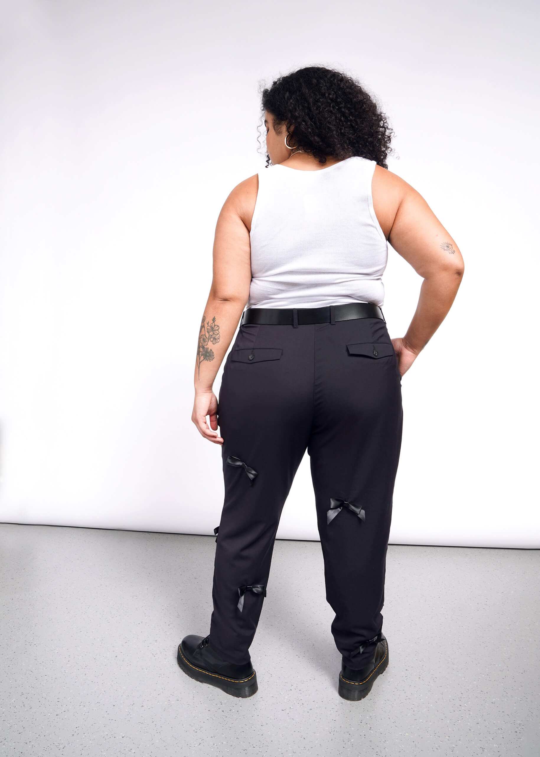 1X=A person with curly hair stands against a white wall wearing a white tank top and The Empower Bows Trouser, ultra-luxe mid-rise black pants adorned with small bows. Tattoos cover their arms, and they complete the look with black shoes.