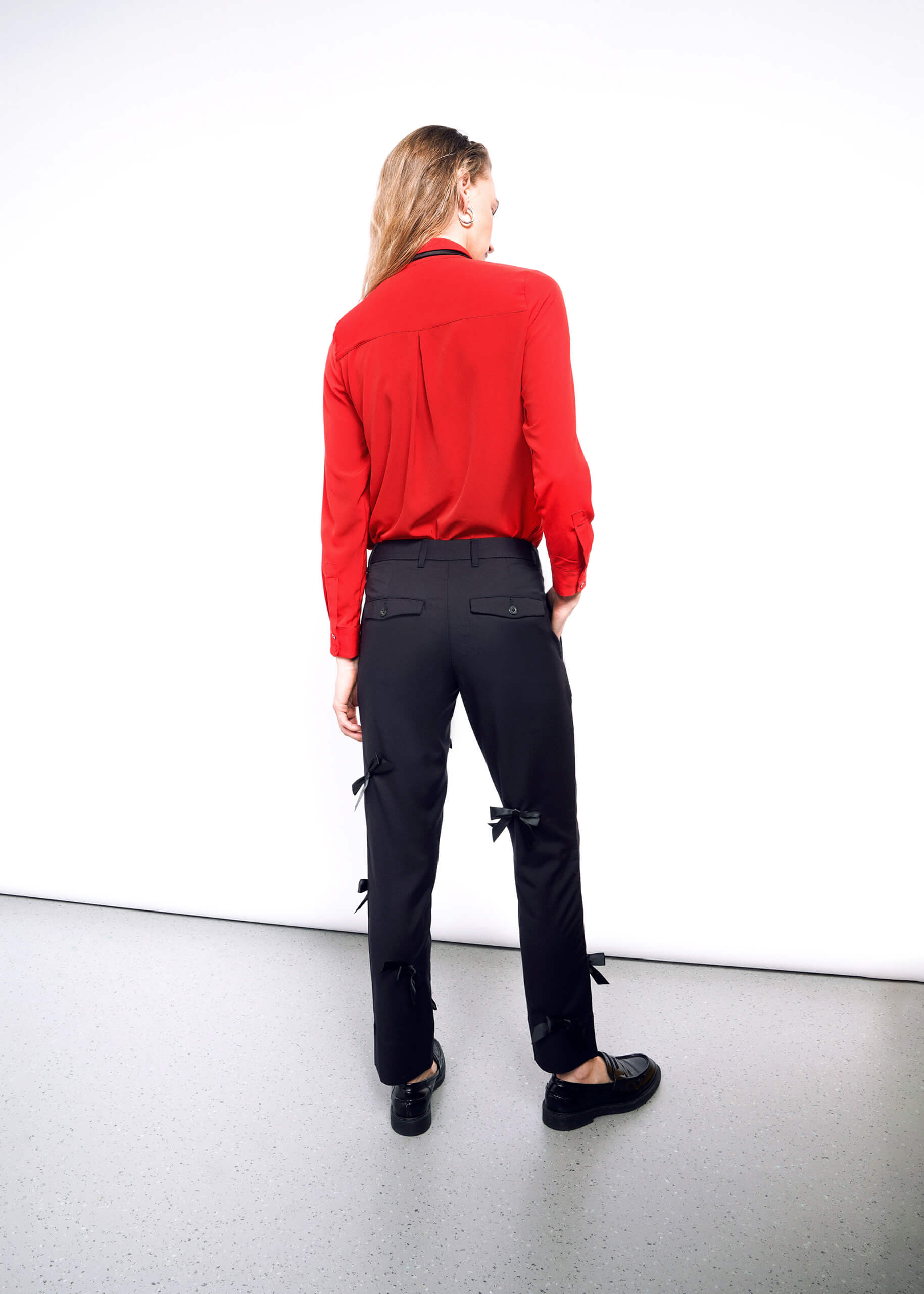 S=Against a white wall, a person wears an ultra-lux red long-sleeve shirt paired with The Empower Bows Trouser, featuring bow details on the sides. Facing away from the camera, they finish their stylish look with sleek black shoes.