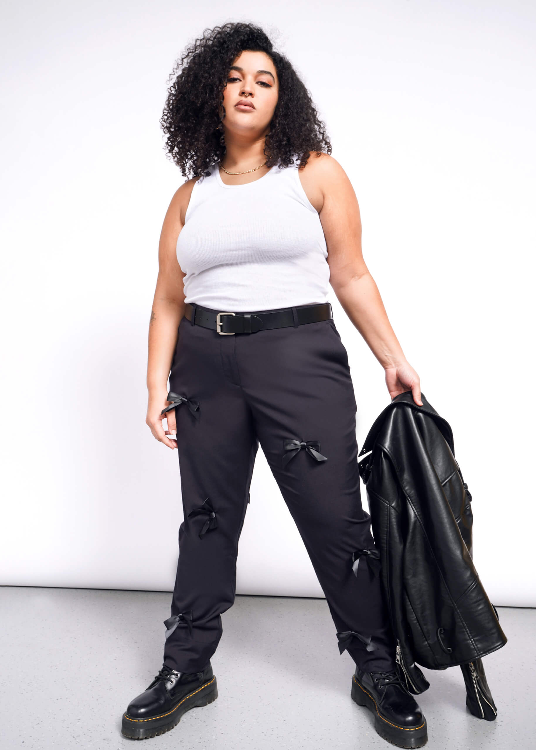 1X= A curly-haired woman in a white tank top and The Empower Bows Trouser stands confidently, holding a leather jacket. Her ultra-lux style is completed with black boots against a plain background.