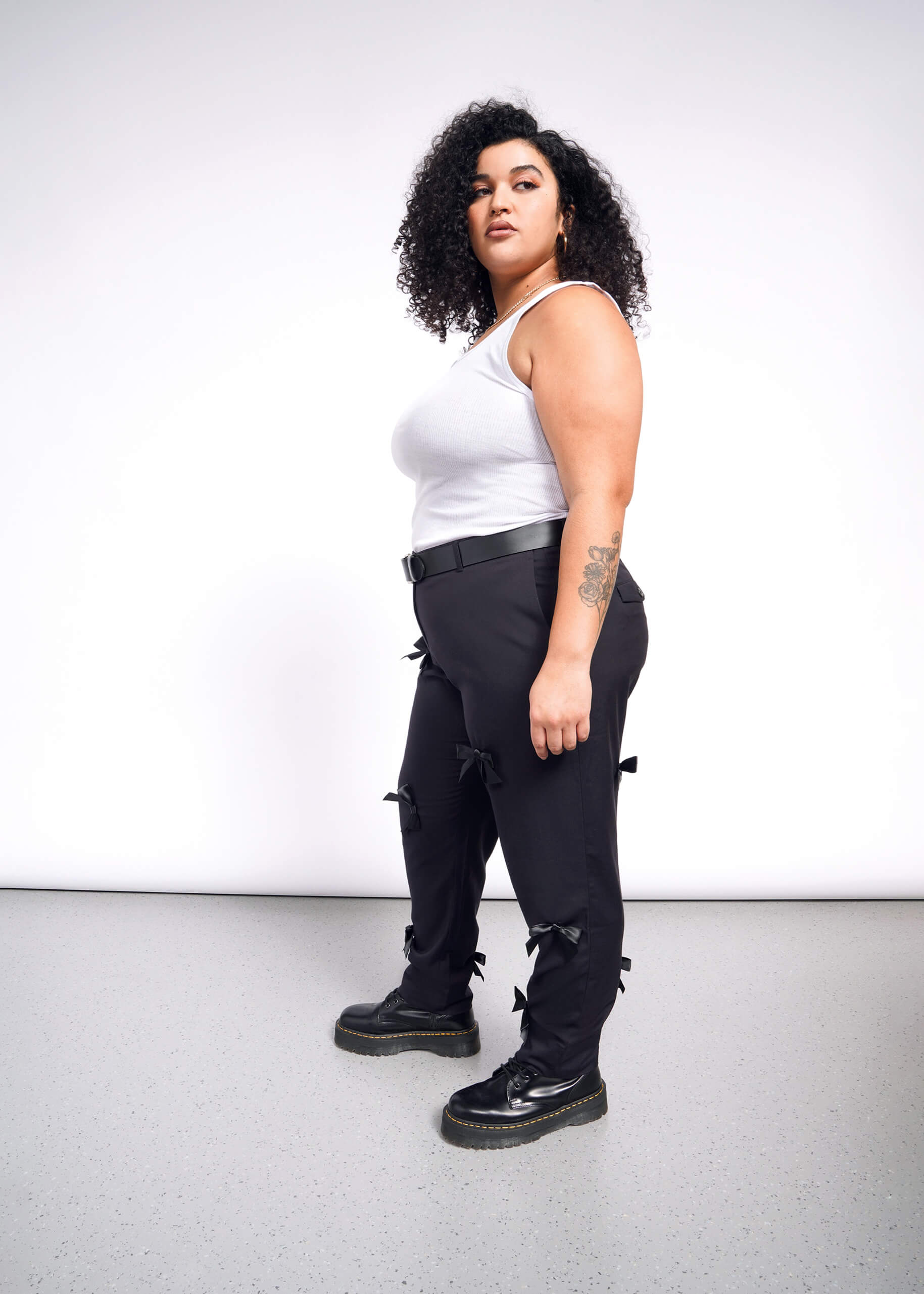 1X=A person with curly hair in a white tank top and The Empower Bows Trouser stands on a speckled floor against a white background. Their right arm features a tattoo, and black shoes complete the chic ensemble.