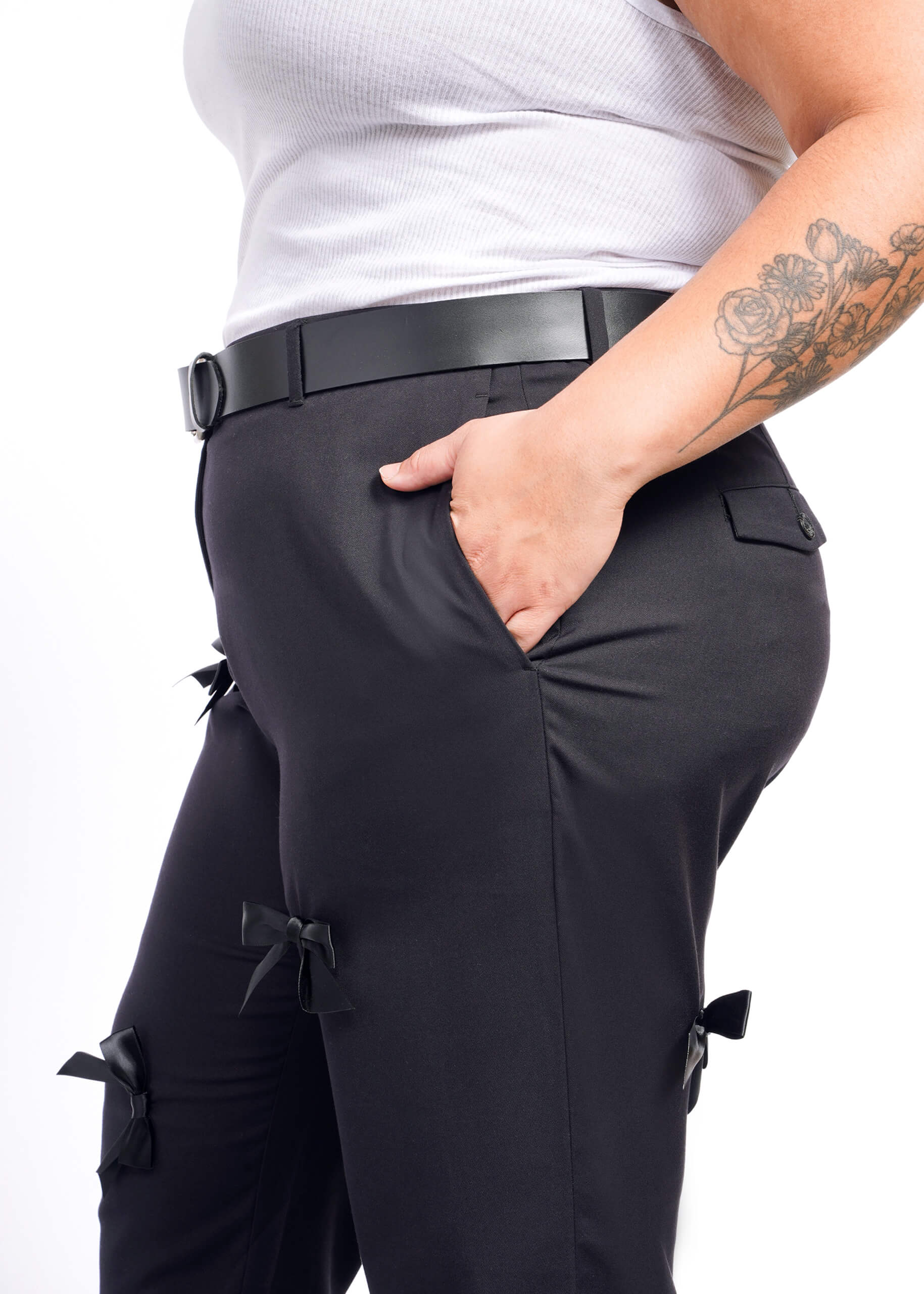 1X= A person wearing The Empower Bows Trouser with a black belt and white tank top is seen against a crisp white background. One hand in their pocket reveals a floral tattoo, exuding an ultra-lux feel.