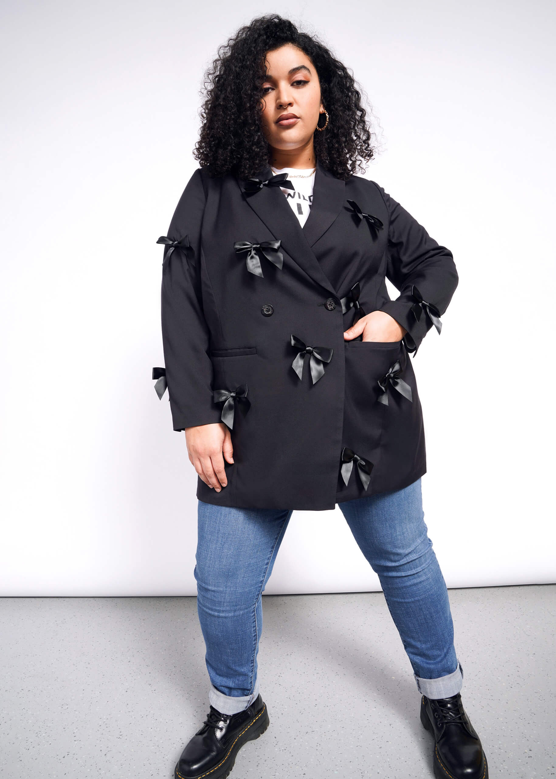 1X= A person with curly hair confidently stands against a white background, wearing The Empower Bows Double Breasted Blazer over a white shirt, paired with blue jeans and black shoes.