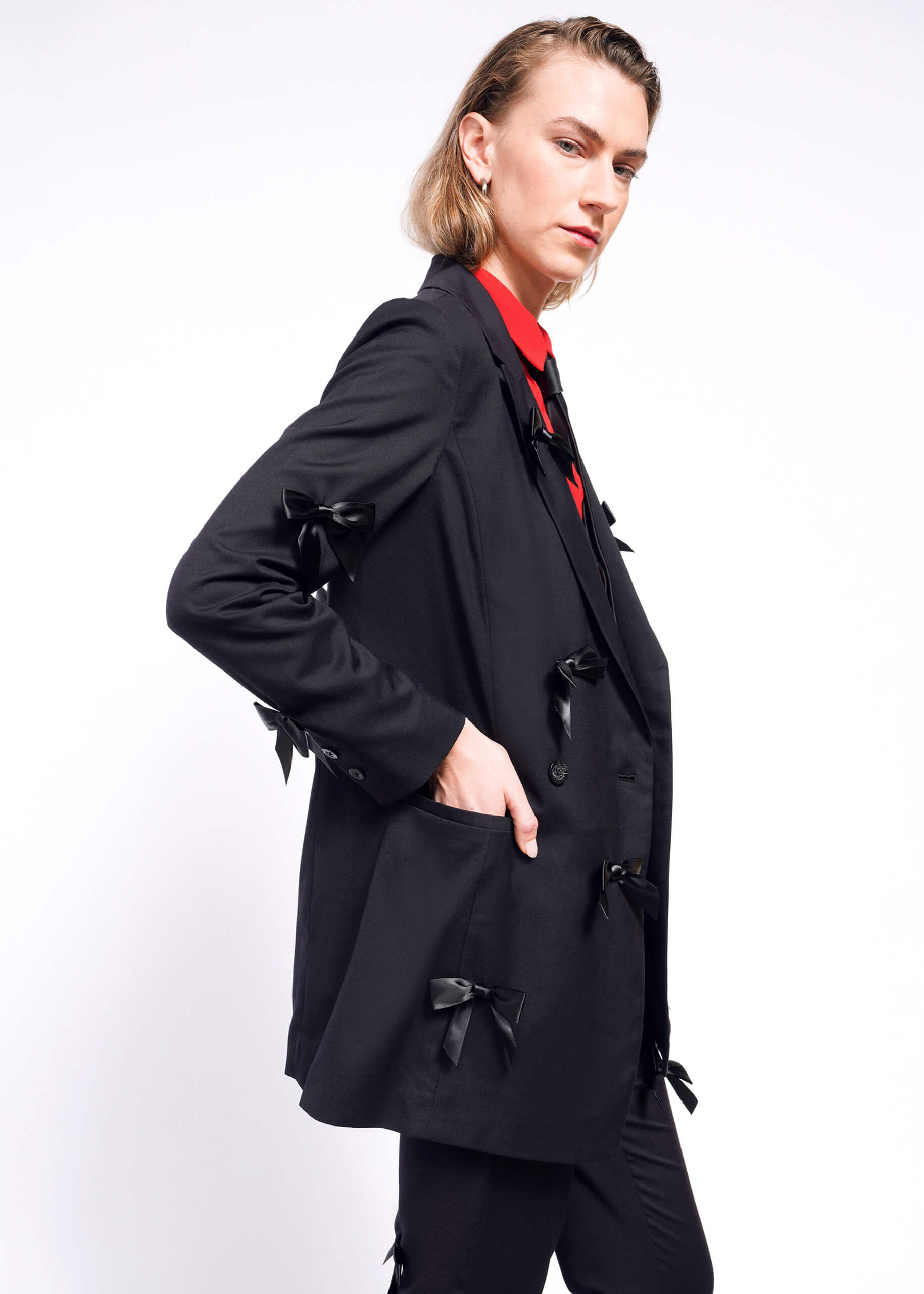 S=Wearing The Empower Bows Double Breasted Blazer, adorned with black bows and a red shirt, a person with shoulder-length hair stands sideways against a white background. With their hand in the blazer pocket, they exude confidence.