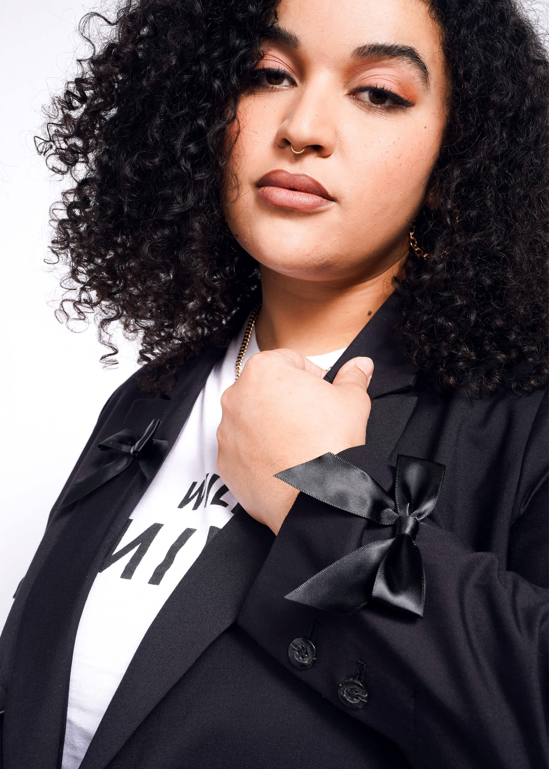 1X=A person with curly hair poses confidently in The Empower Bows Double Breasted Blazer, featuring tailored oversized design and bow details on the sleeves, over a white T-shirt against a white background.