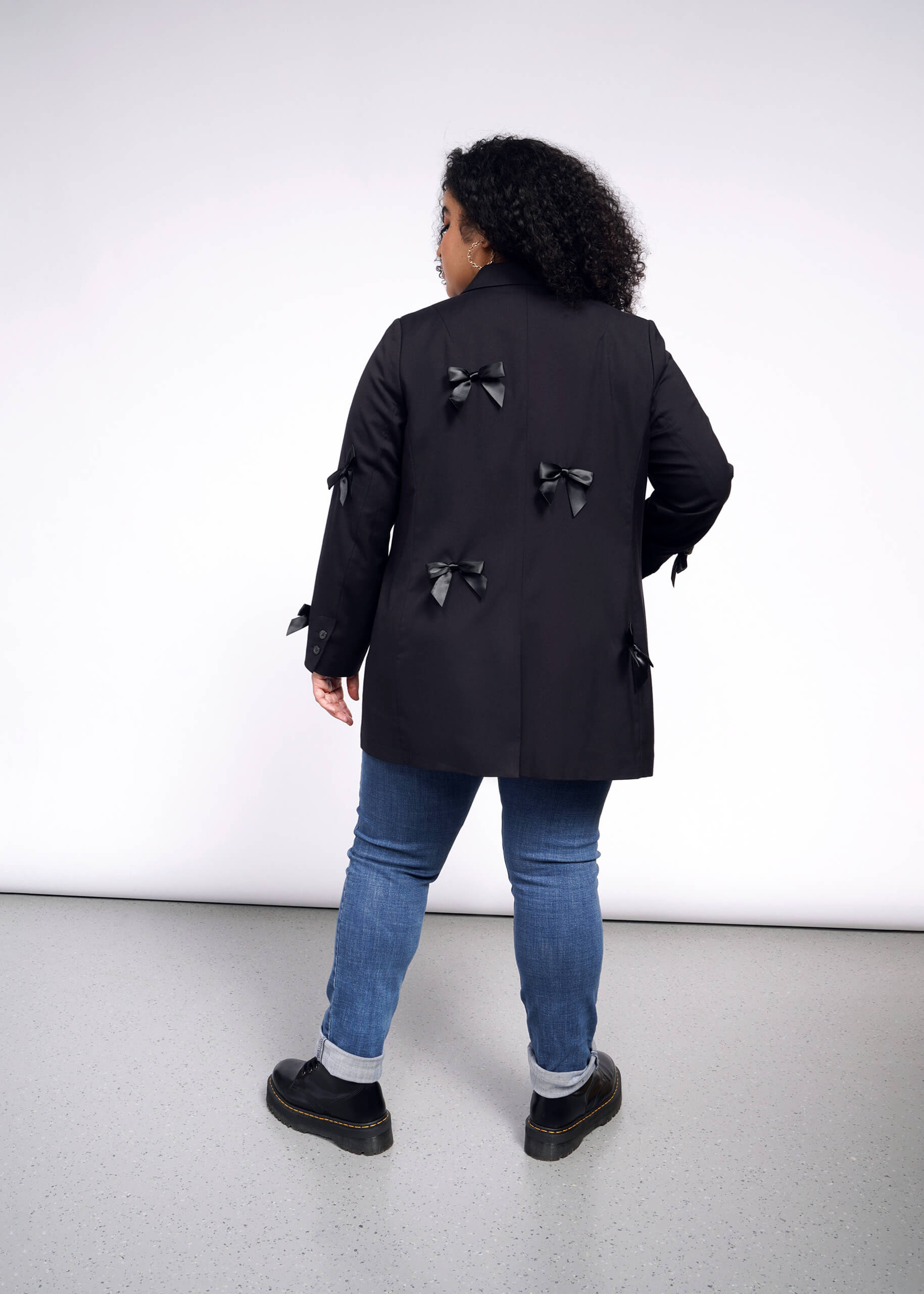 1X=A person with curly hair faces away, wearing an oversized black coat adorned with multiple bows on the back and sleeves. They pair it with blue jeans and black shoes, standing on a light gray floor against a white background. The ensemble flows elegantly like The Empower Bows Double Breasted Blazer.