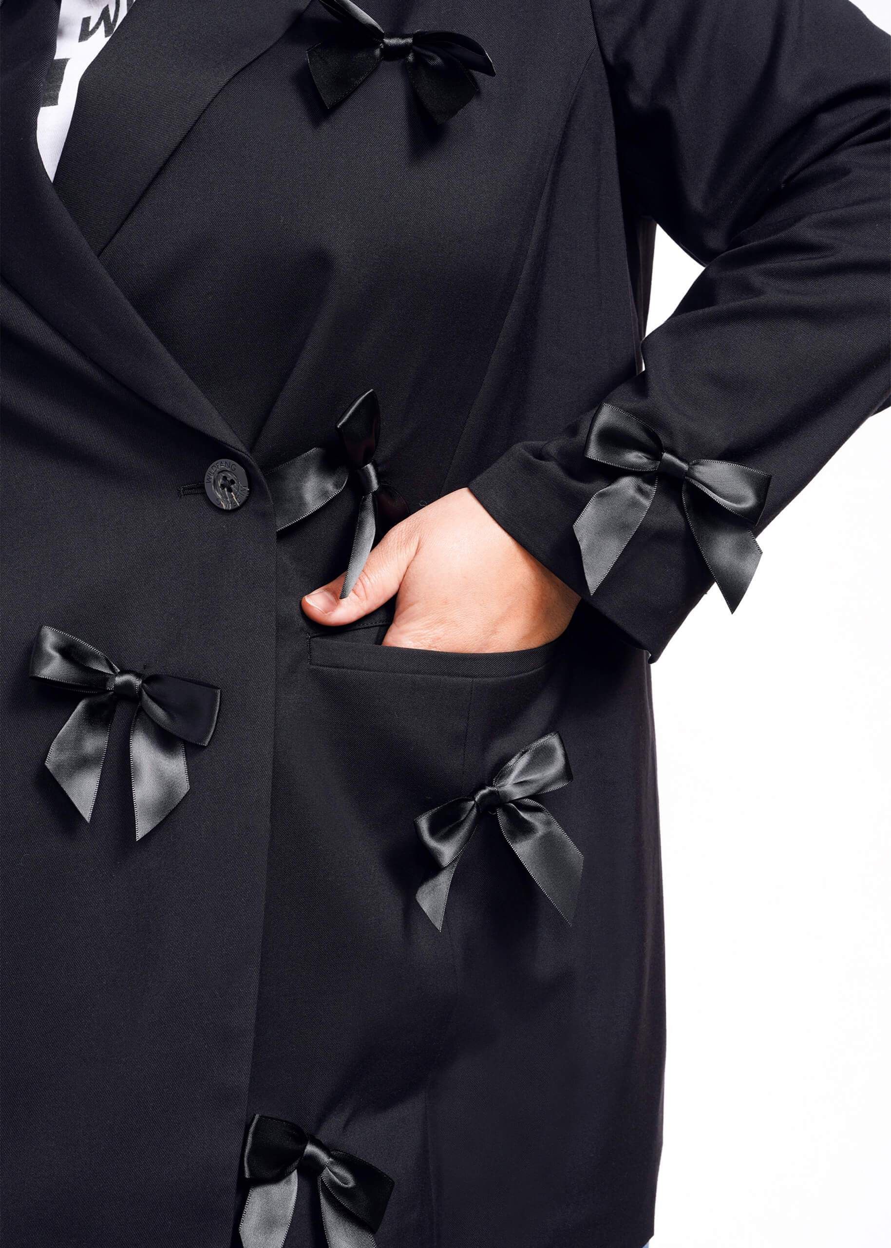 1X= A person wearing The Empower Bows Double Breasted Blazer is photographed against a white background. The tailored oversized coat features black satin bows and drapes elegantly, with their left hand resting in one of its pockets.