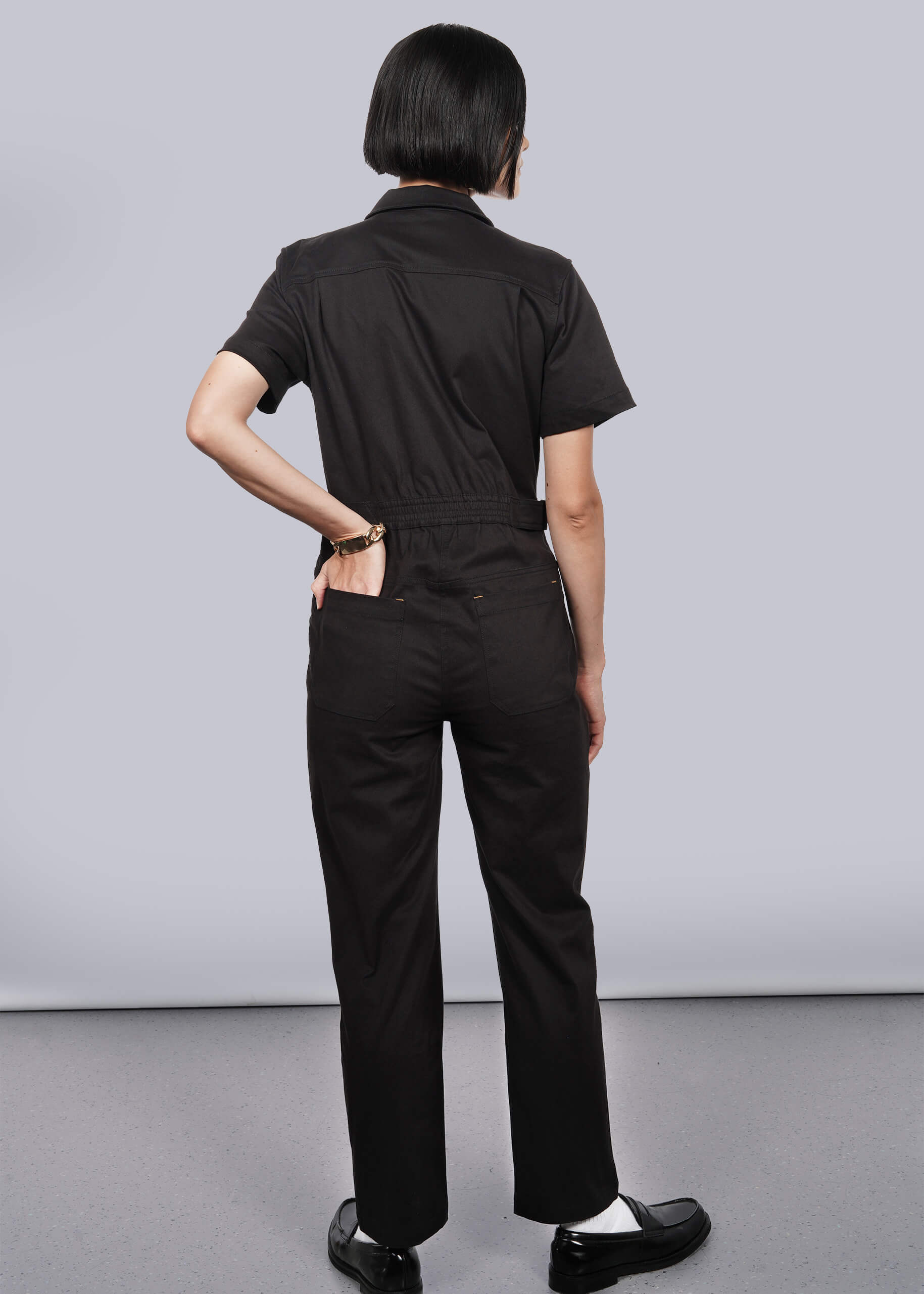 S= A person with short dark hair, wearing The Essential High Waisted Coverall in black—featuring a high-waisted fit and deep pockets—stands against a plain light background, hand on hip, paired with black shoes and white socks.