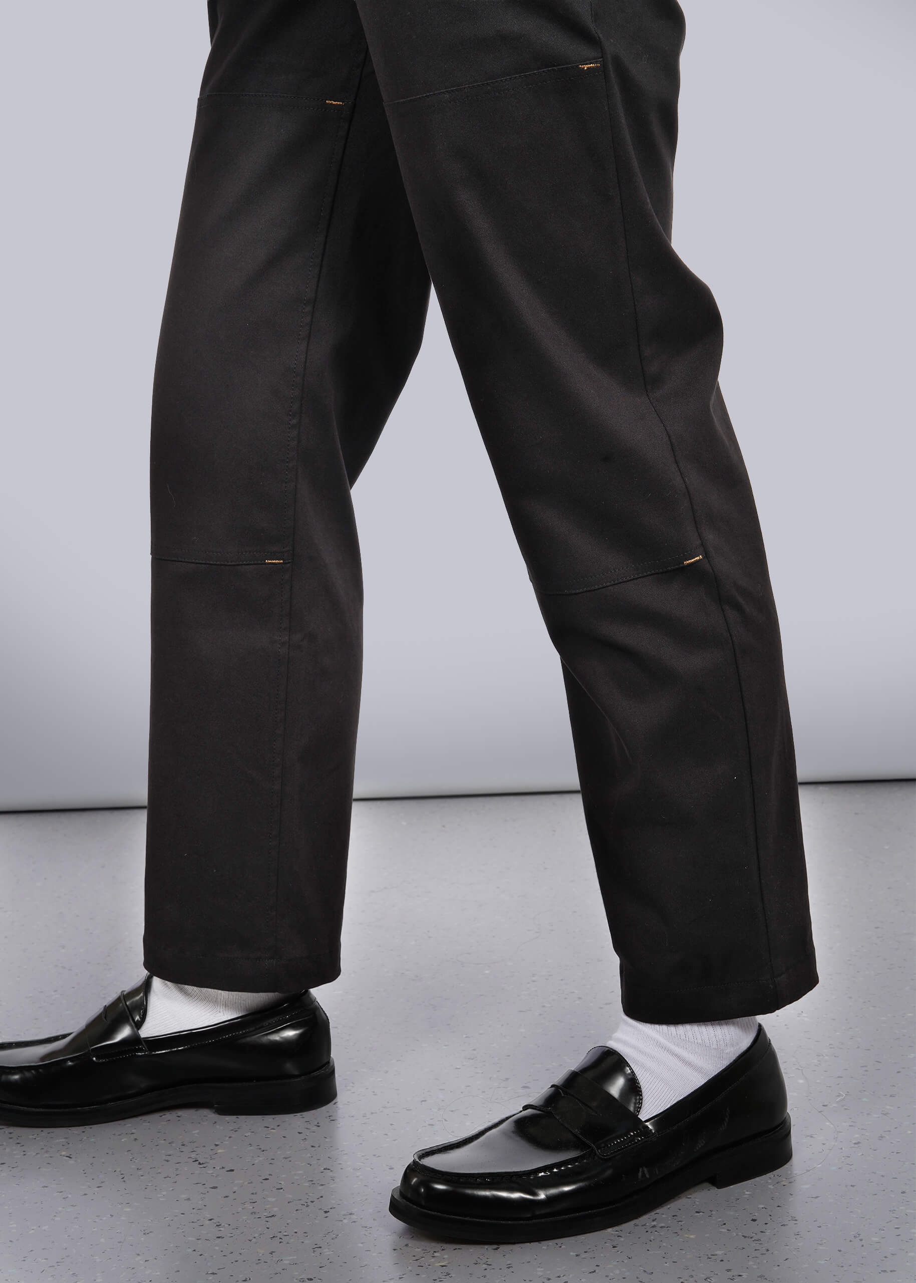 S=Wearing The Essential High Waisted Coverall, a person stands on a gray speckled floor with only their lower legs and feet—clad in white socks and shiny black loafers—visible against a plain white background.