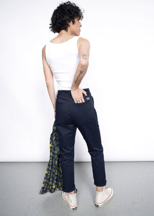S=A person with short curly hair, wearing a white tank top, The Essential Slim Crop Pant in a tailored mid-rise fit, and white sneakers faces away holding a patterned jacket. Tattoos show on their left arm as they stand on a gray floor against a white background.