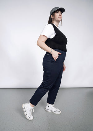 1X=A person stands sideways against a plain background, wearing a gray cap, white t-shirt, black knit vest, and The Essential Slim Crop Pant in navy for a tailored mid-rise fit. White sneakers complete the look as they gaze slightly upward with one hand in their pocket.