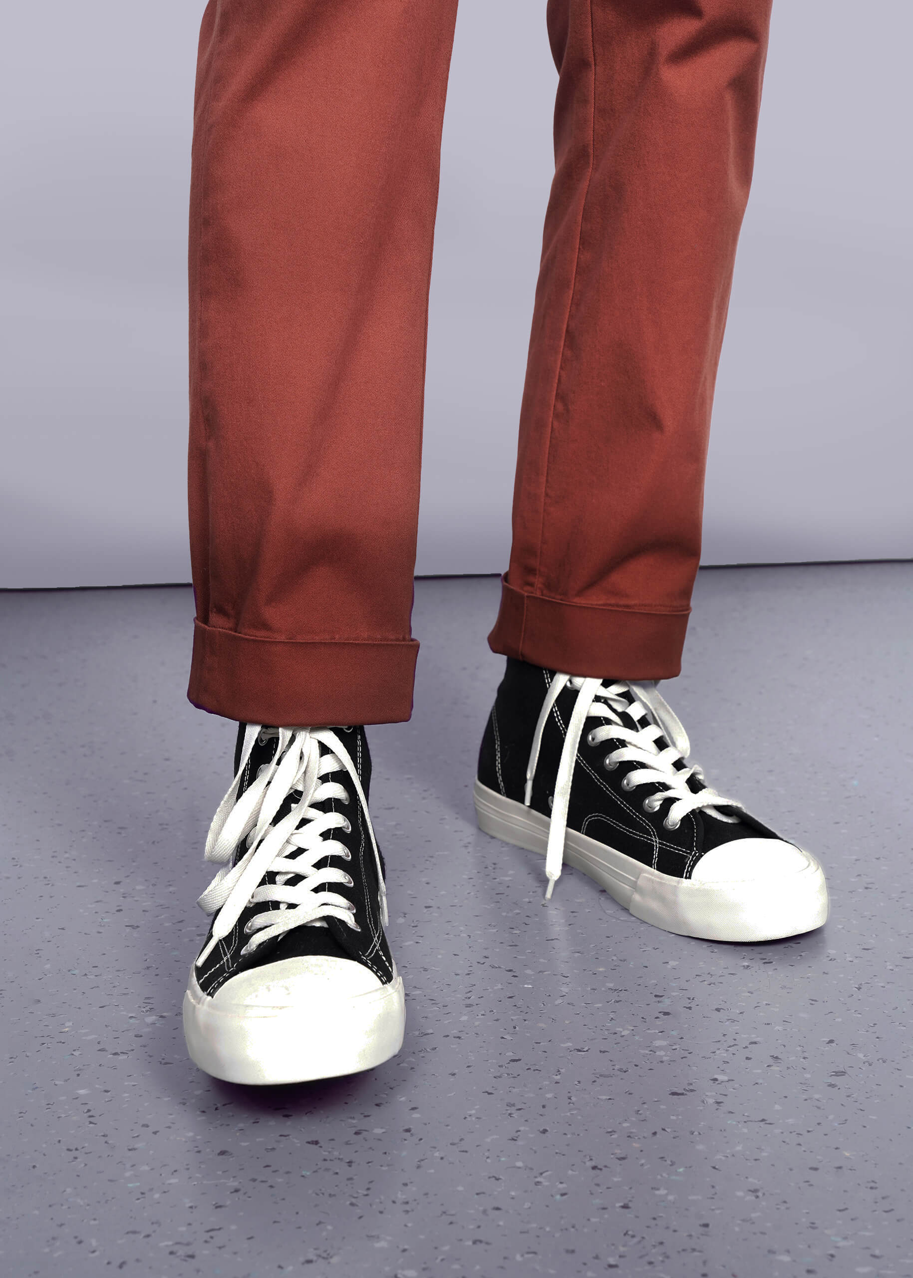 S=Wearing The Essential Trouser in a relaxed rust hue—timeless wardrobe staples—paired with black high-top sneakers featuring white soles and laces, standing on a light gray speckled floor against a plain background.
