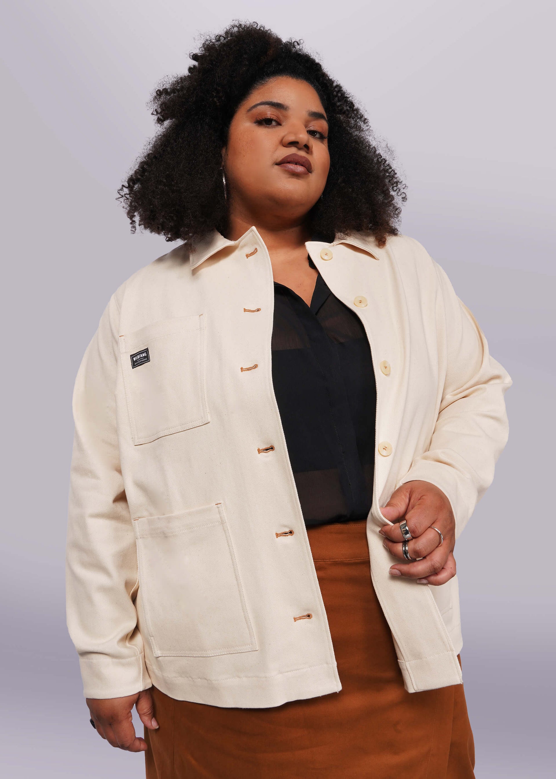 1X=A person with curly hair poses confidently, wearing The Essential Denim Utility Jacket over a black top and a brown skirt. The cream-colored jacket features deep pockets and a label, set against a plain, light background.