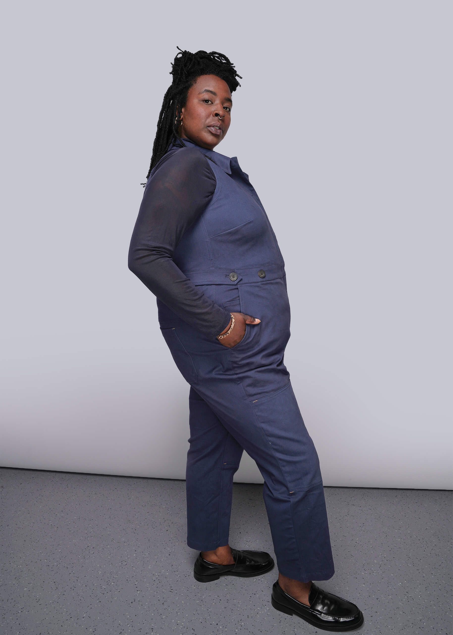 1X=Person wearing a blue coverall with hand in side pocket against a plain background