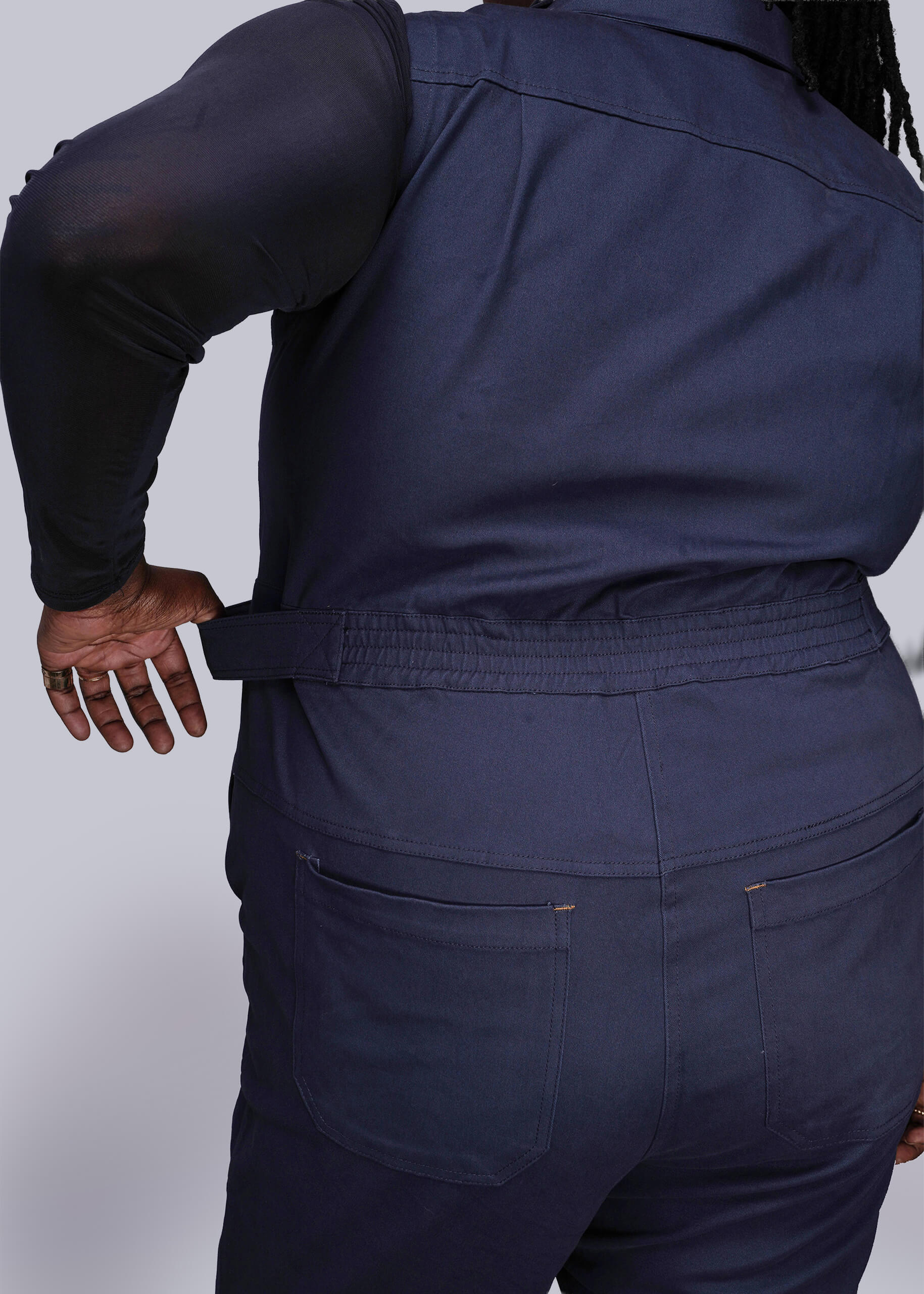 1X=Back of person wearing a navy blue jumpsuit holding waistband on a plain background