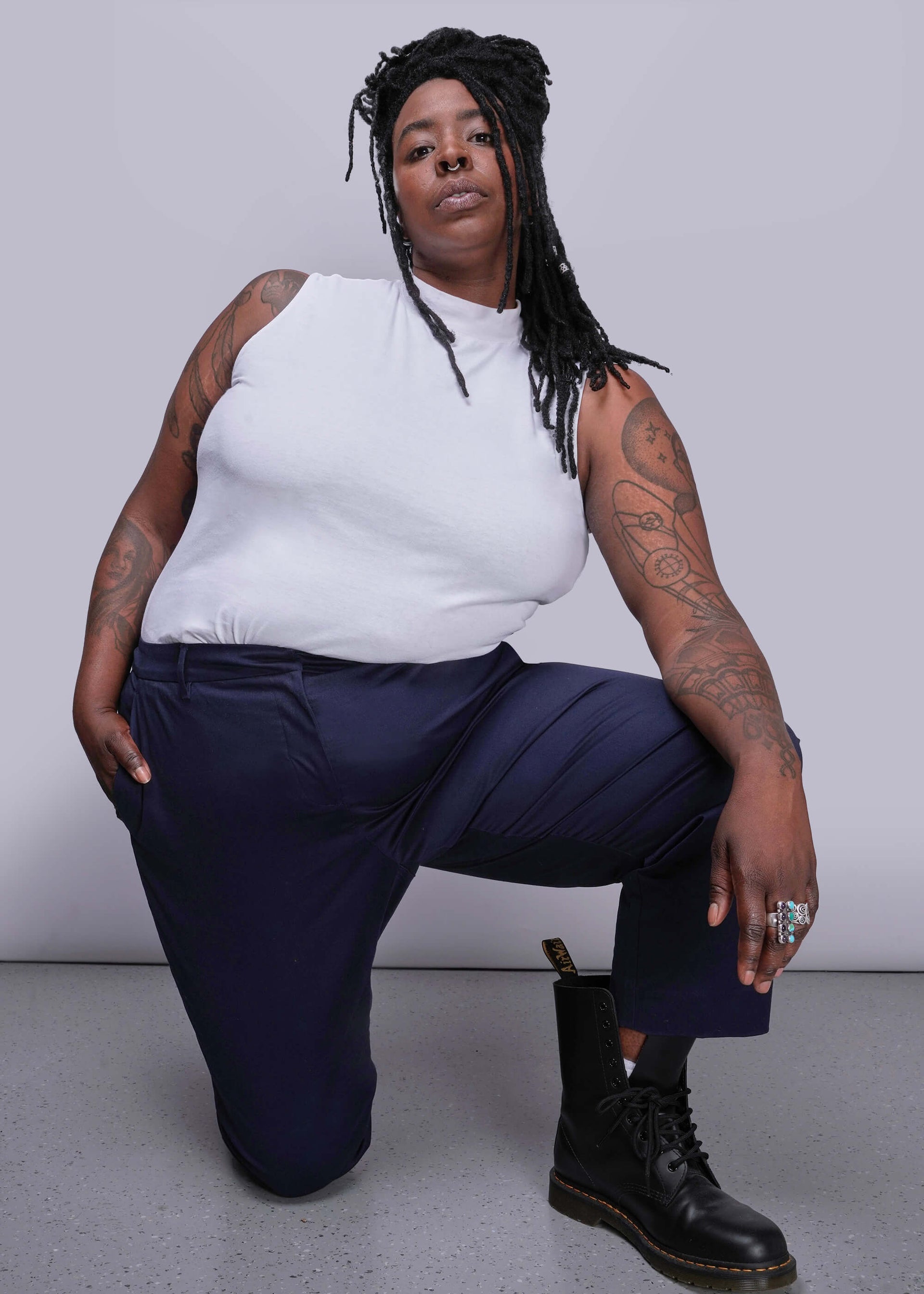 1X=Person wearing a white sleeveless top and navy pants, sitting on the floor against a plain background.