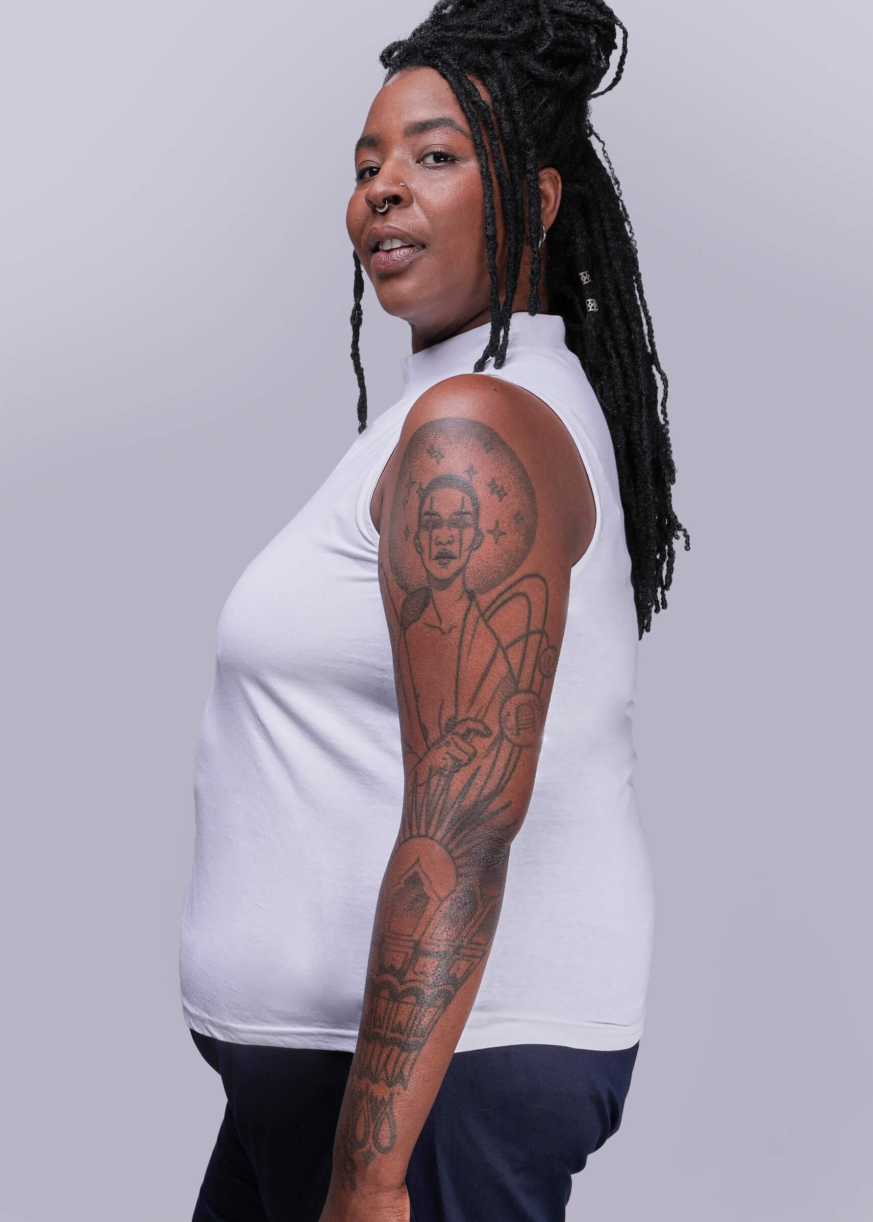 1X=Person wearing a white sleeveless top with tattoos on arms against a gray background
