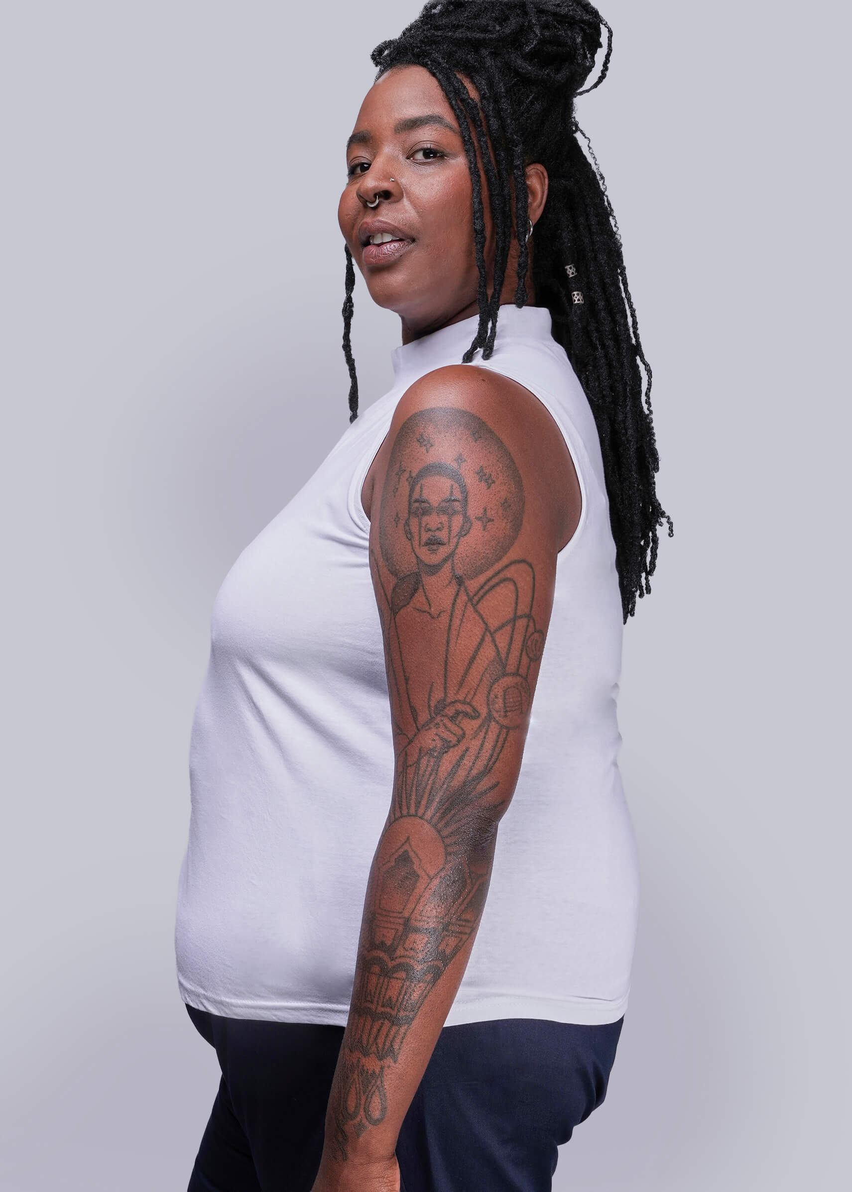 1X=Person wearing a white sleeveless top with tattoos on arms against a gray background