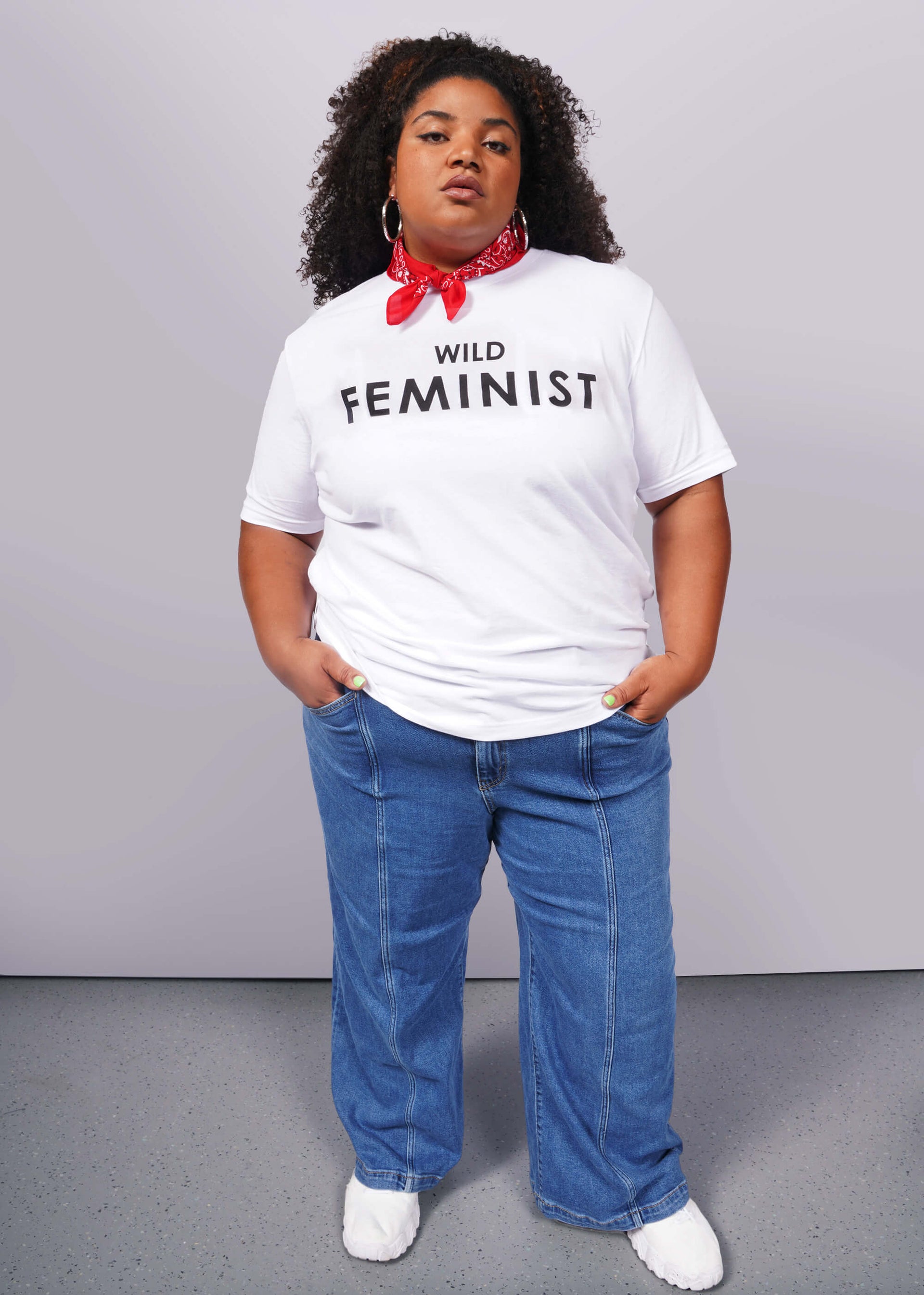 1X= A confident person wears The Wild Feminist® Tee, a relaxed unisex fit white shirt, paired with a red bandana, blue jeans, and white shoes. The plain background emphasizes their commitment to unity and advocacy.