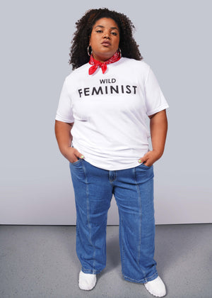 1X= A confident person wears The Wild Feminist® Tee, a relaxed unisex fit white shirt, paired with a red bandana, blue jeans, and white shoes. The plain background emphasizes their commitment to unity and advocacy.