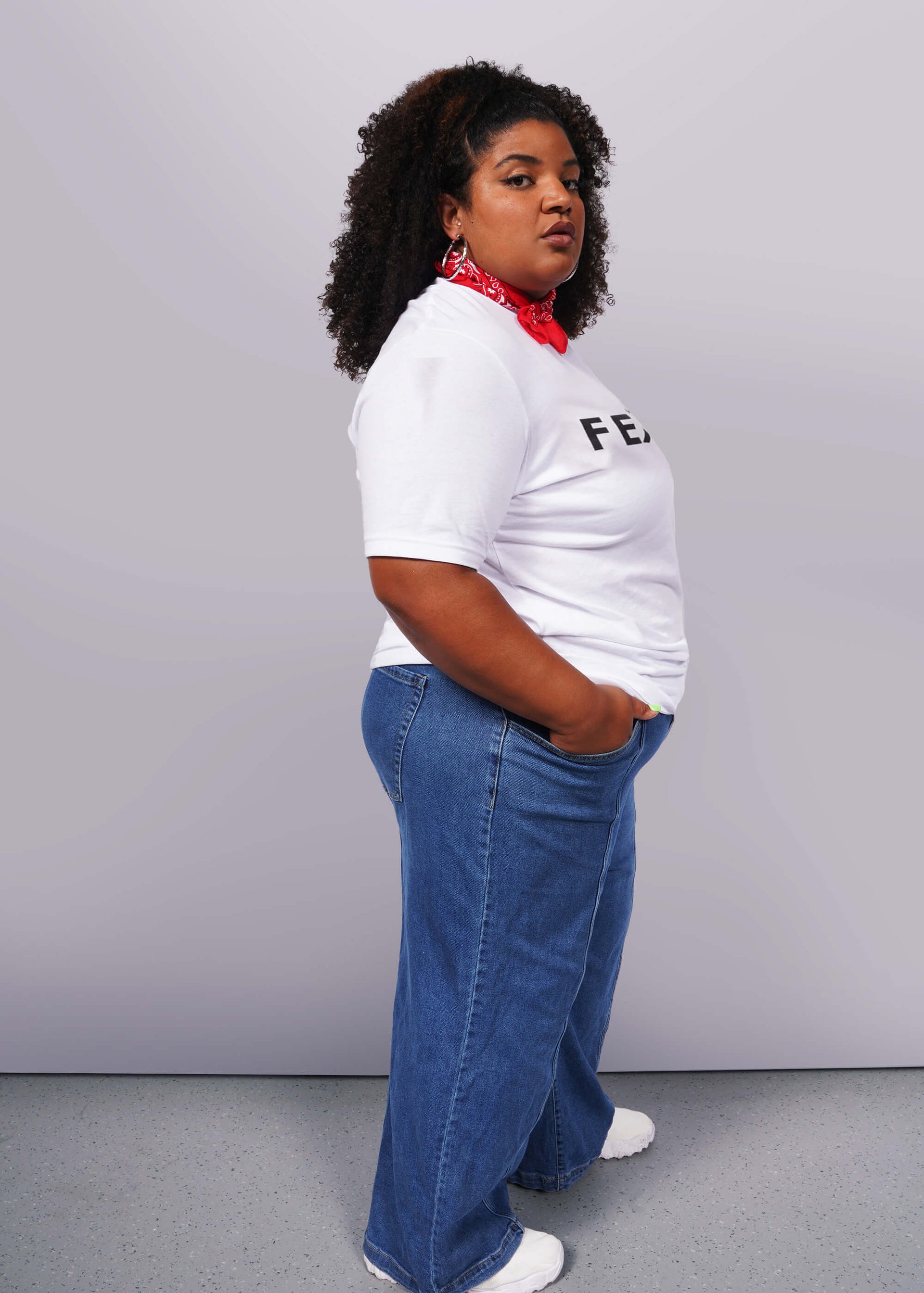 1X=A person with curly hair confidently poses against a gray background, wearing a relaxed unisex fit. They sport The Wild Feminist® Tee, blue jeans, white sneakers, and a red bandana around their neck. One hand in their pocket, they embody unity.