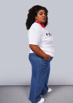 1X=A person with curly hair confidently poses against a gray background, wearing a relaxed unisex fit. They sport The Wild Feminist® Tee, blue jeans, white sneakers, and a red bandana around their neck. One hand in their pocket, they embody unity.