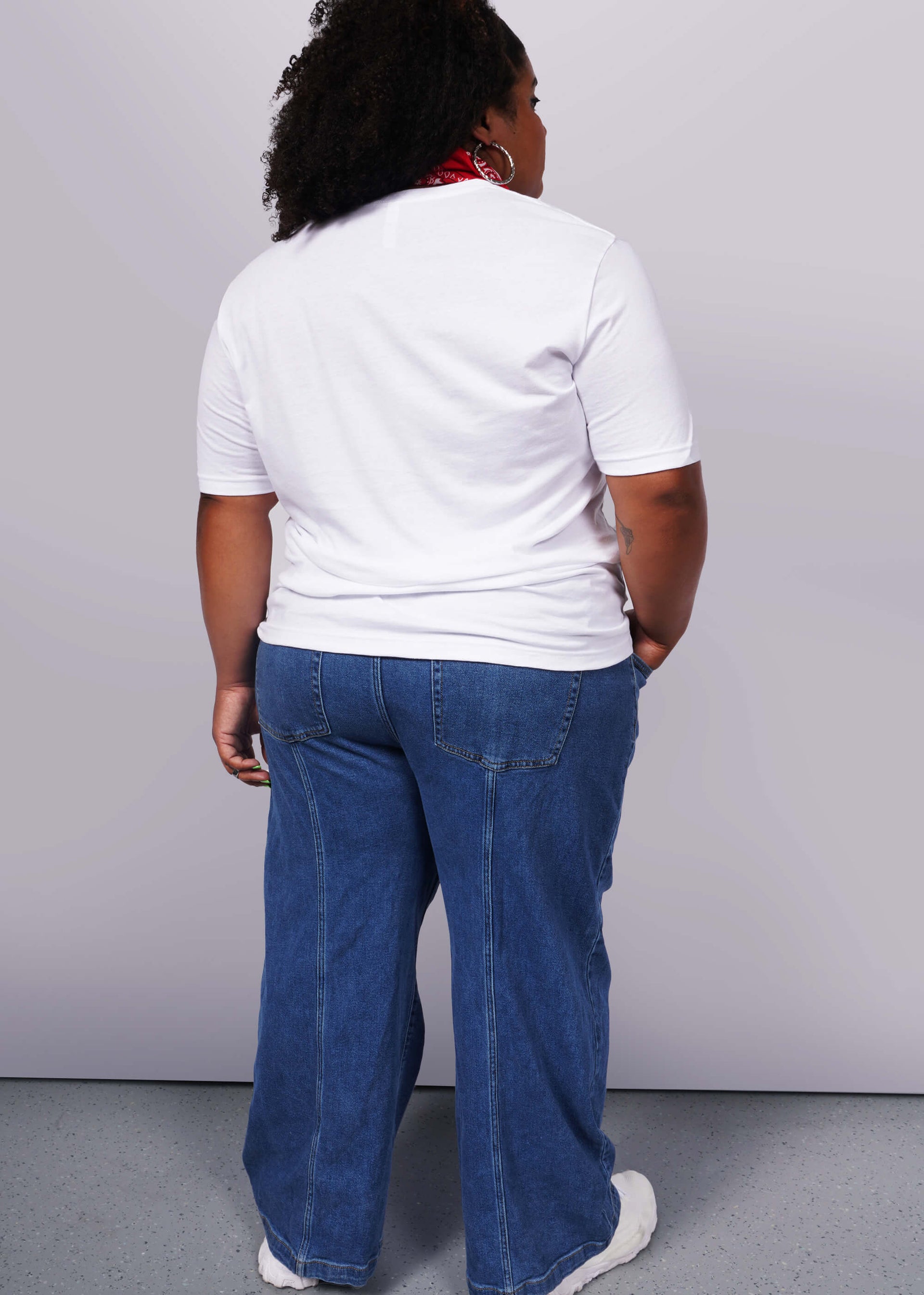 1X= A person with curly hair and relaxed poise stands with their back to the camera in a unisex white Wild Feminist® Tee and blue jeans, accented by a red scarf. The plain light gray background highlights their effortless style.
