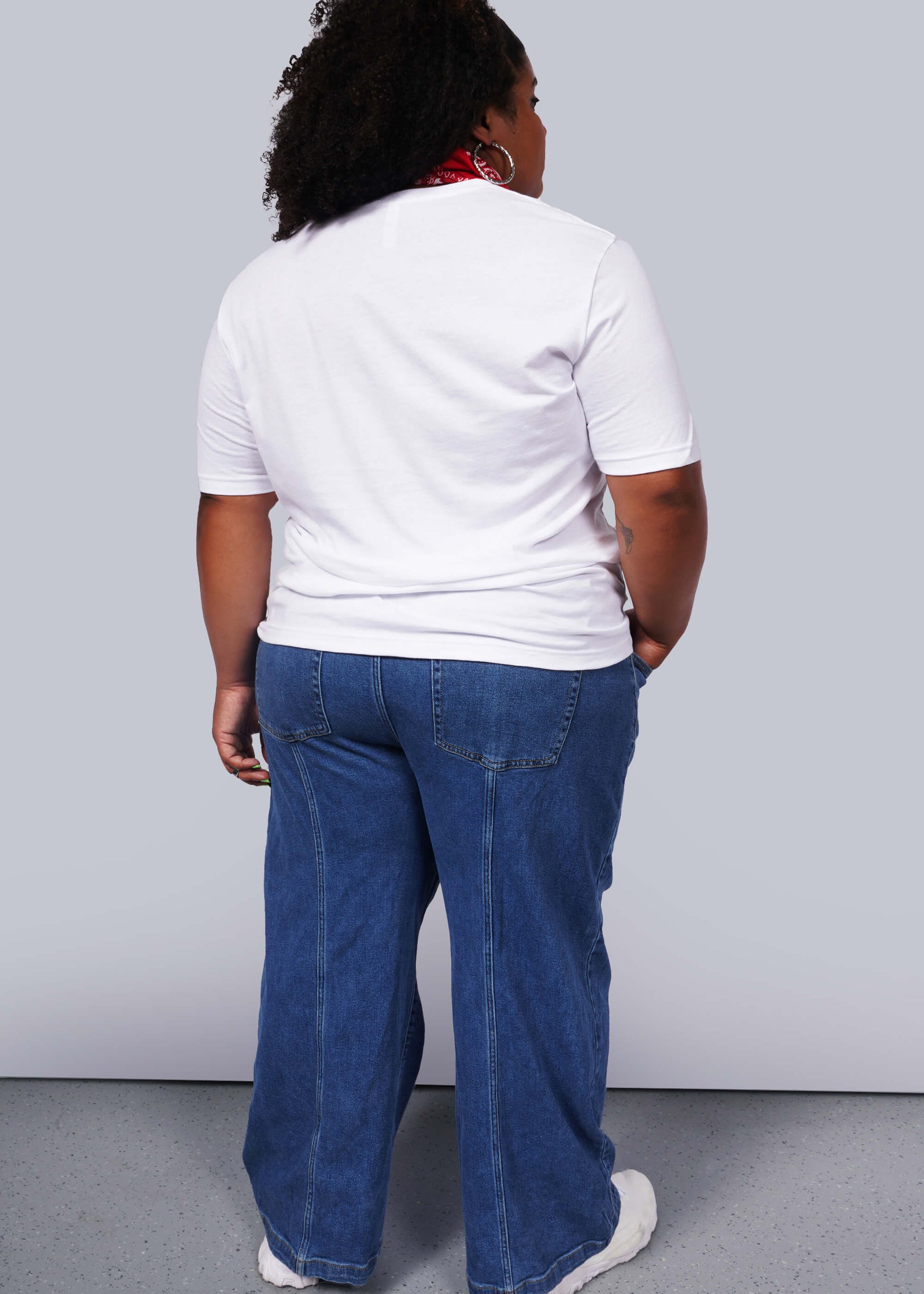 1X= A person with curly hair and relaxed poise stands with their back to the camera in a unisex white Wild Feminist® Tee and blue jeans, accented by a red scarf. The plain light gray background highlights their effortless style.