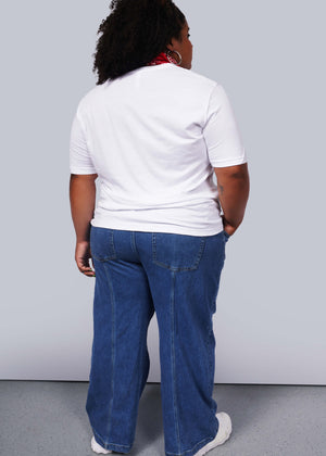1X= A person with curly hair and relaxed poise stands with their back to the camera in a unisex white Wild Feminist® Tee and blue jeans, accented by a red scarf. The plain light gray background highlights their effortless style.