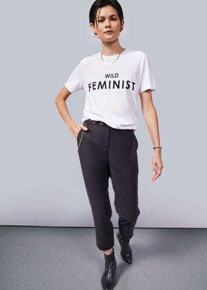 S= A person with short dark hair wears The Wild Feminist® Tee, black pants, and black ankle boots, embodying a relaxed unisex fit. Standing confidently against a plain background with one hand in their pocket, they inspire unity.