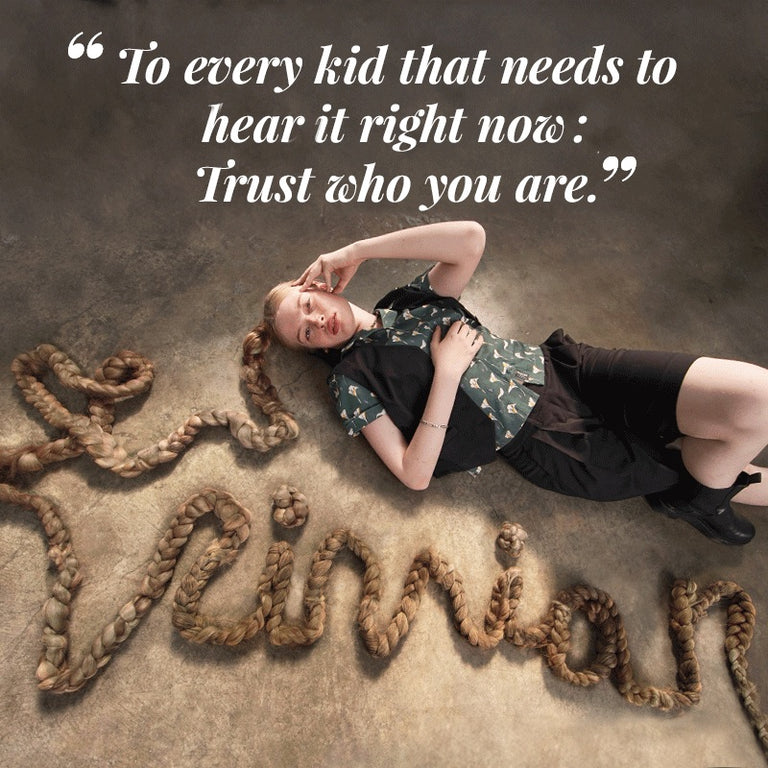 Vivian Wilson lays on floor with one leg crossed over the other, her hair is braided and is styled very long so that it spells out VIVIAN on the floor in cursive on front of her. She's wearing The Empower Vest in black, The Everyday Camp Button Up, and The Empower Wide Leg Short in Black. Text overlay on top of image is a quote from Vivian. It reads "To every kid that needs to hear it right now: trust who you are."