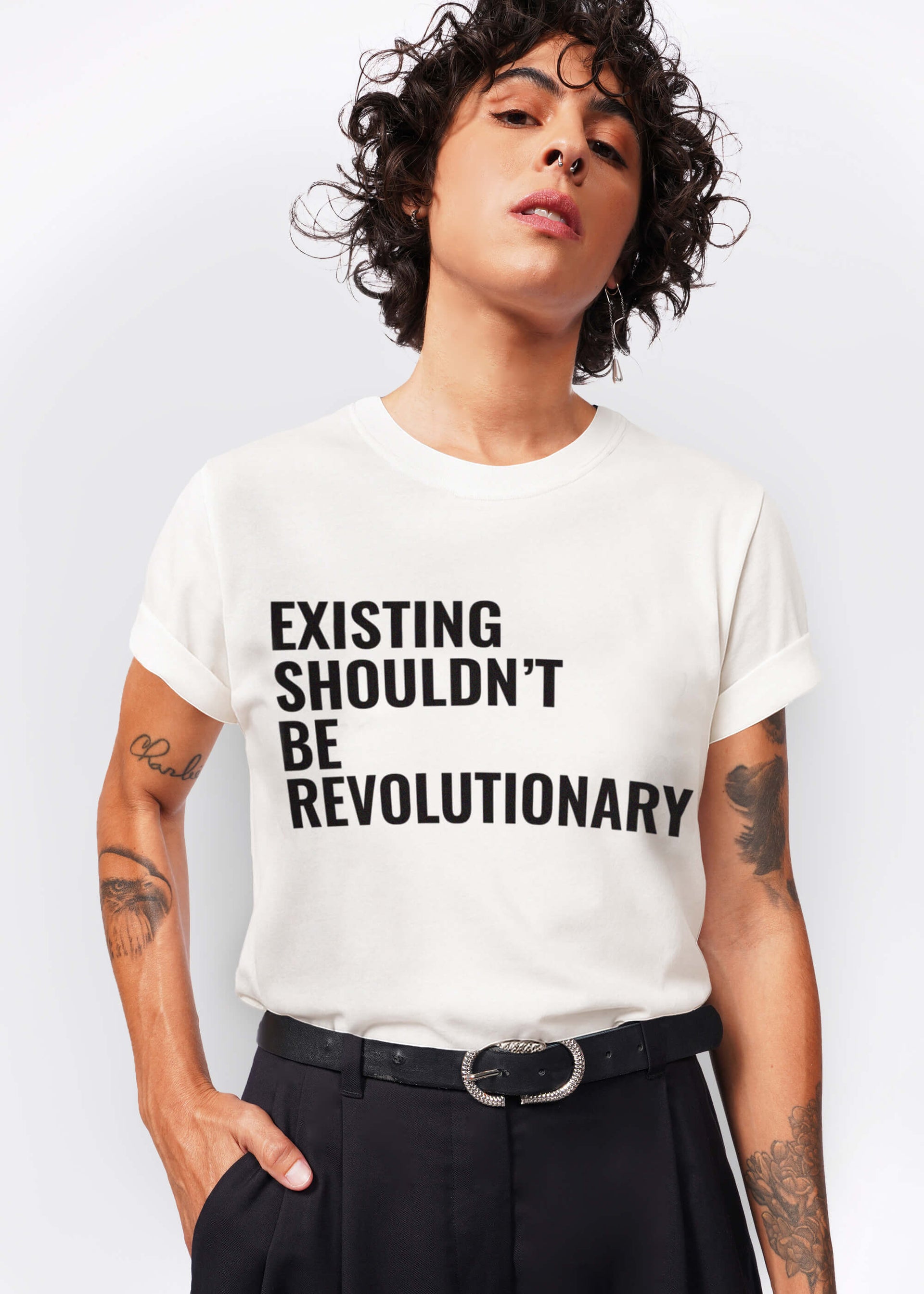 S= A person with short curly hair wears the VIVIAN WILSON FOR WILDFANG TEE—a white shirt boldly stating “EXISTING SHOULDN’T BE REVOLUTIONARY”—standing confidently, hands in pockets, proud to support Trans lives matter.