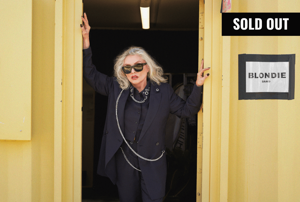 Debbie Harry stands in doorway of yellow metal building with BLONDIE sign on it. She's wearing sunglasses and a suit from the Debbie Harry x Wildfang collection. Black banner in top right corner reads SOLD OUT