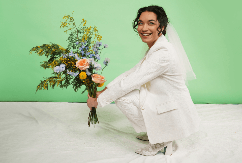 Woman with curly black hair pulled into bun crouches down in front of green and white background holding bouquet and wearing the David's Bridal and Wildfang collab suit in a white lace fabric.