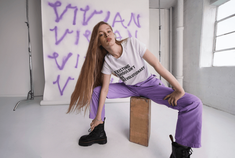 Vivian Wilson sits on wood block leaning over with hand on ankle, her blonde hair draped over her shoulder, wearing The Essential High Waisted Coverall in Lilac, the Vivian Wilson for Wildfang Tee, and Doc Marten black boots. Behind her, a white backdrop features her name spray painted in purple multiple times.