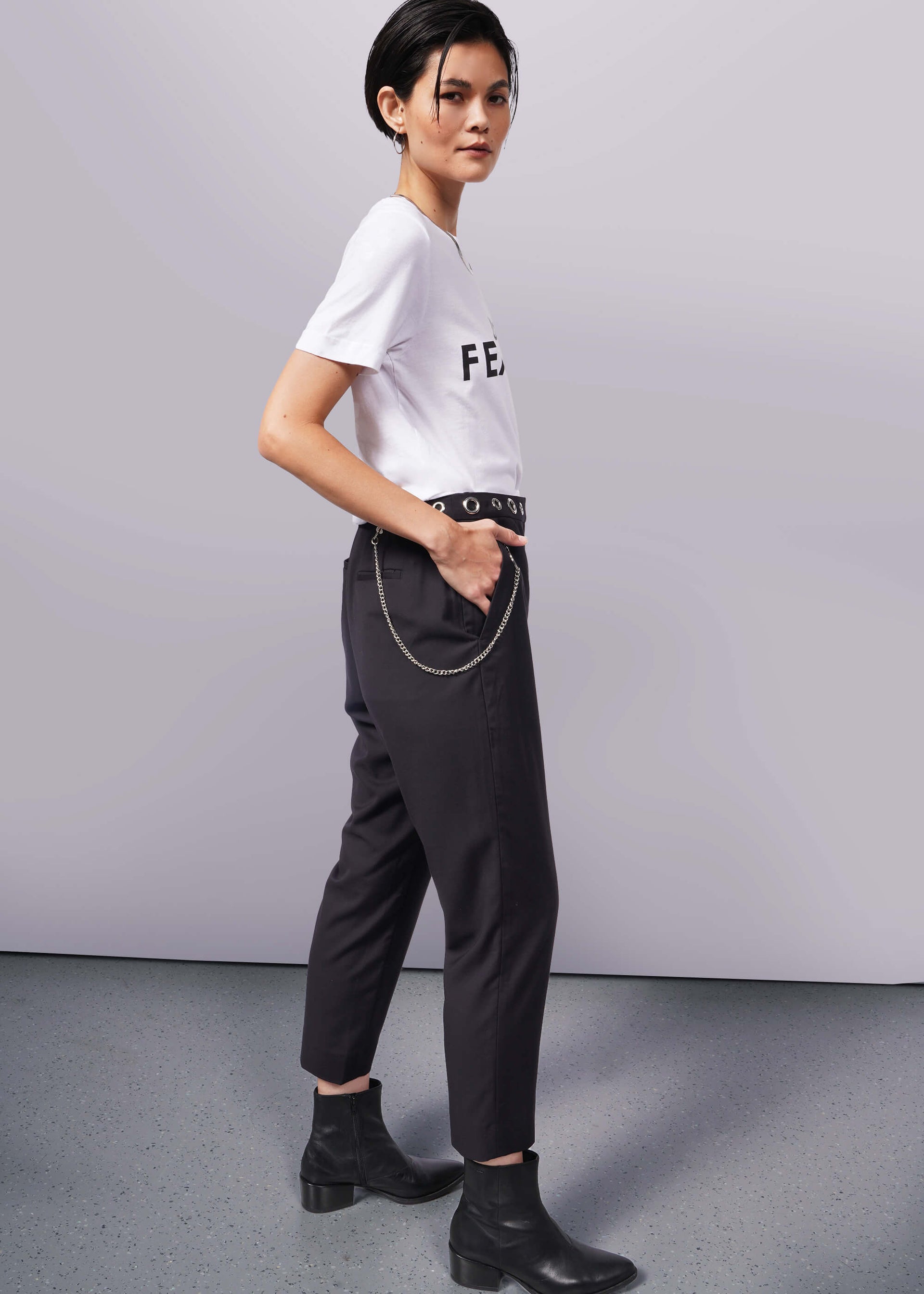 S=A person with short dark hair stands in profile against a white background, wearing The Wild Feminist® Tee—a relaxed unisex fit—paired with dark pants featuring a chain accessory and black boots. One hand rests casually in their pocket, exuding effortless style.