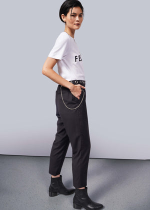 S=A person with short dark hair stands in profile against a white background, wearing The Wild Feminist® Tee—a relaxed unisex fit—paired with dark pants featuring a chain accessory and black boots. One hand rests casually in their pocket, exuding effortless style.