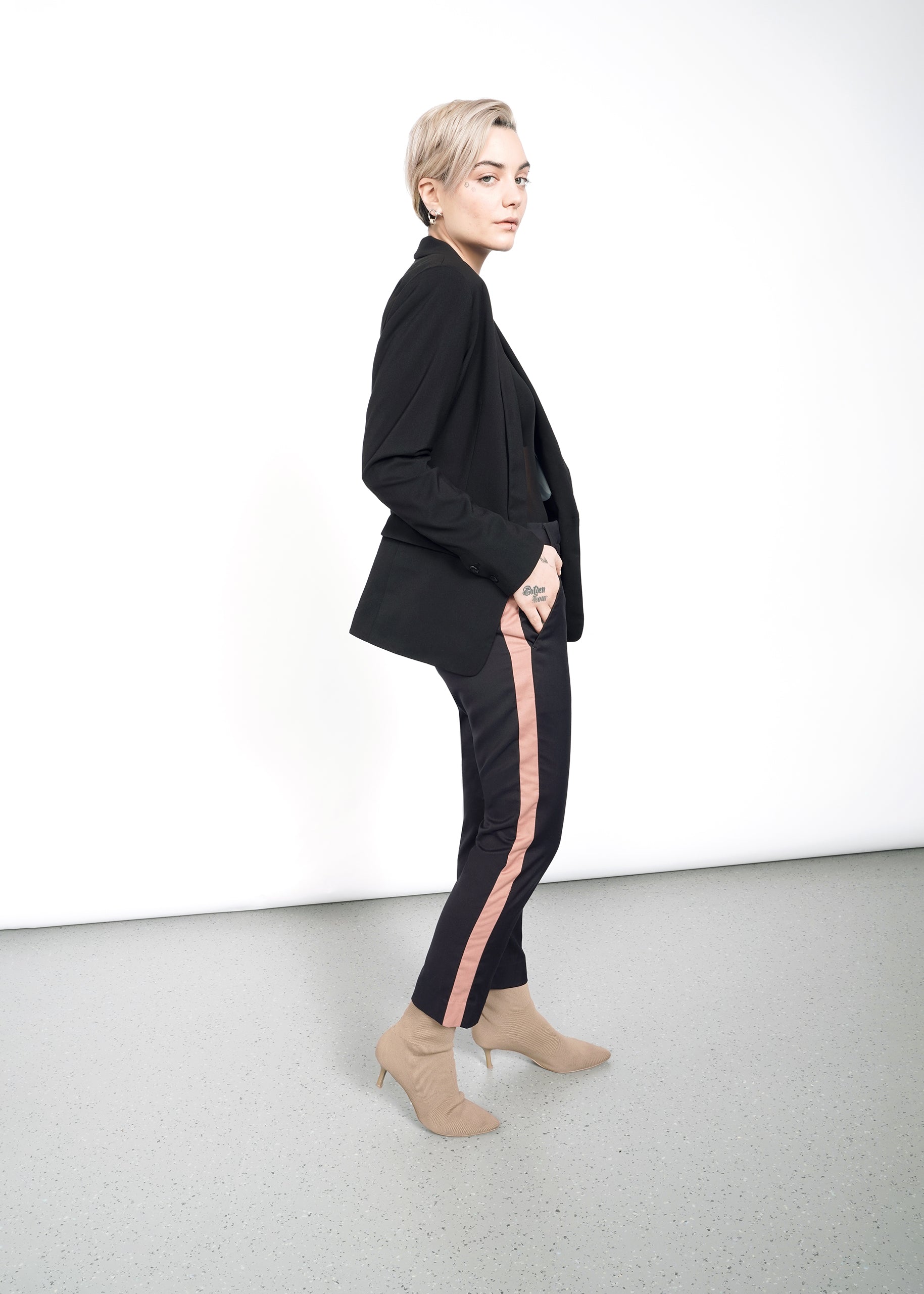 S=A person with short hair wears The Empower Tux Blazer in black, featuring a pink stripe detail, paired with beige ankle boots. They stand against a white wall, flaunting deep pockets on the timeless blazer and showcasing a hand tattoo.