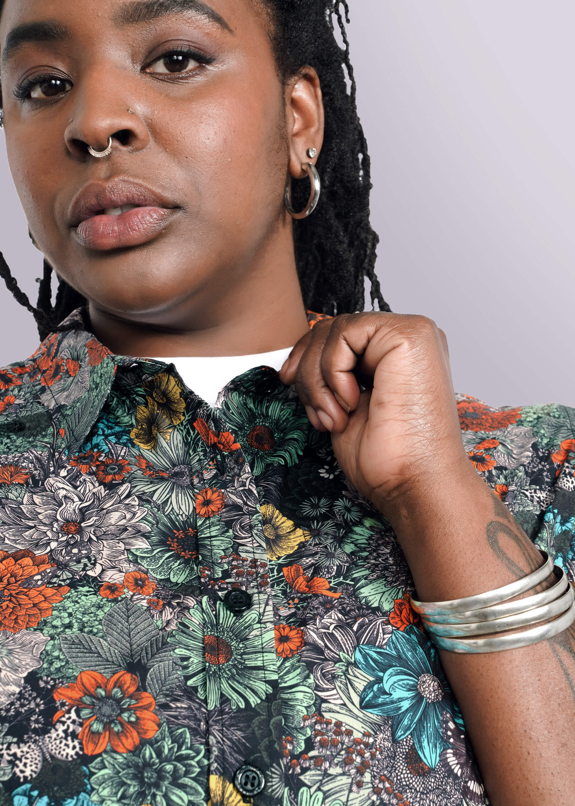 1X=Wearing hoop earrings, a nose ring, and silver bangles, a person with long braided hair gently adjusts the classic collar of The Essential Button Up—a colorful floral shirt with a tailored fit. Made from 100% cotton, the vibrant design stands out on a crisp white background.
