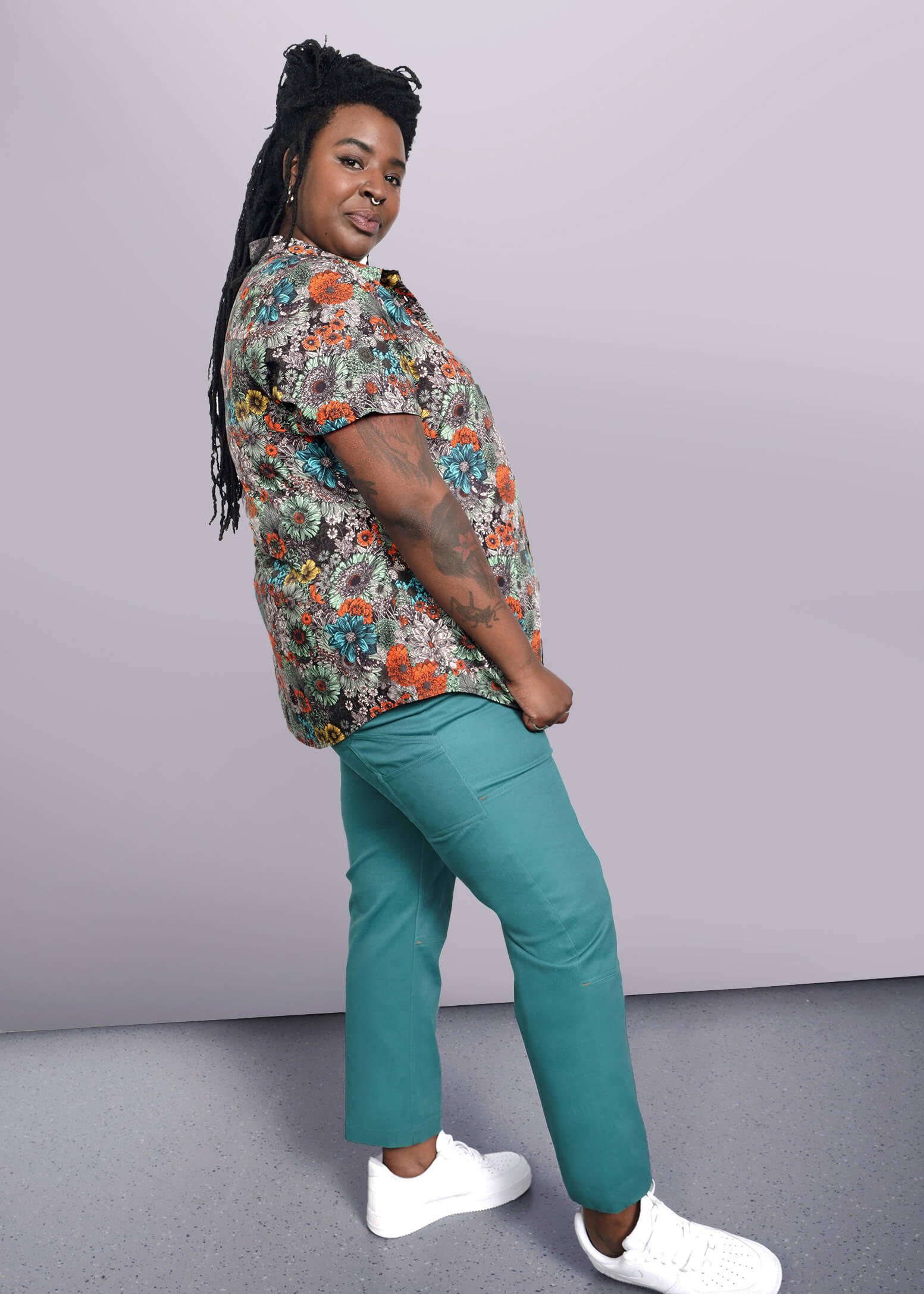 1X=A person with long braided hair stands smiling in a side profile, wearing The Essential Button Up featuring a classic collar. Paired with teal pants and white sneakers, the tailored fit enhances their elegance against the minimalist white background.