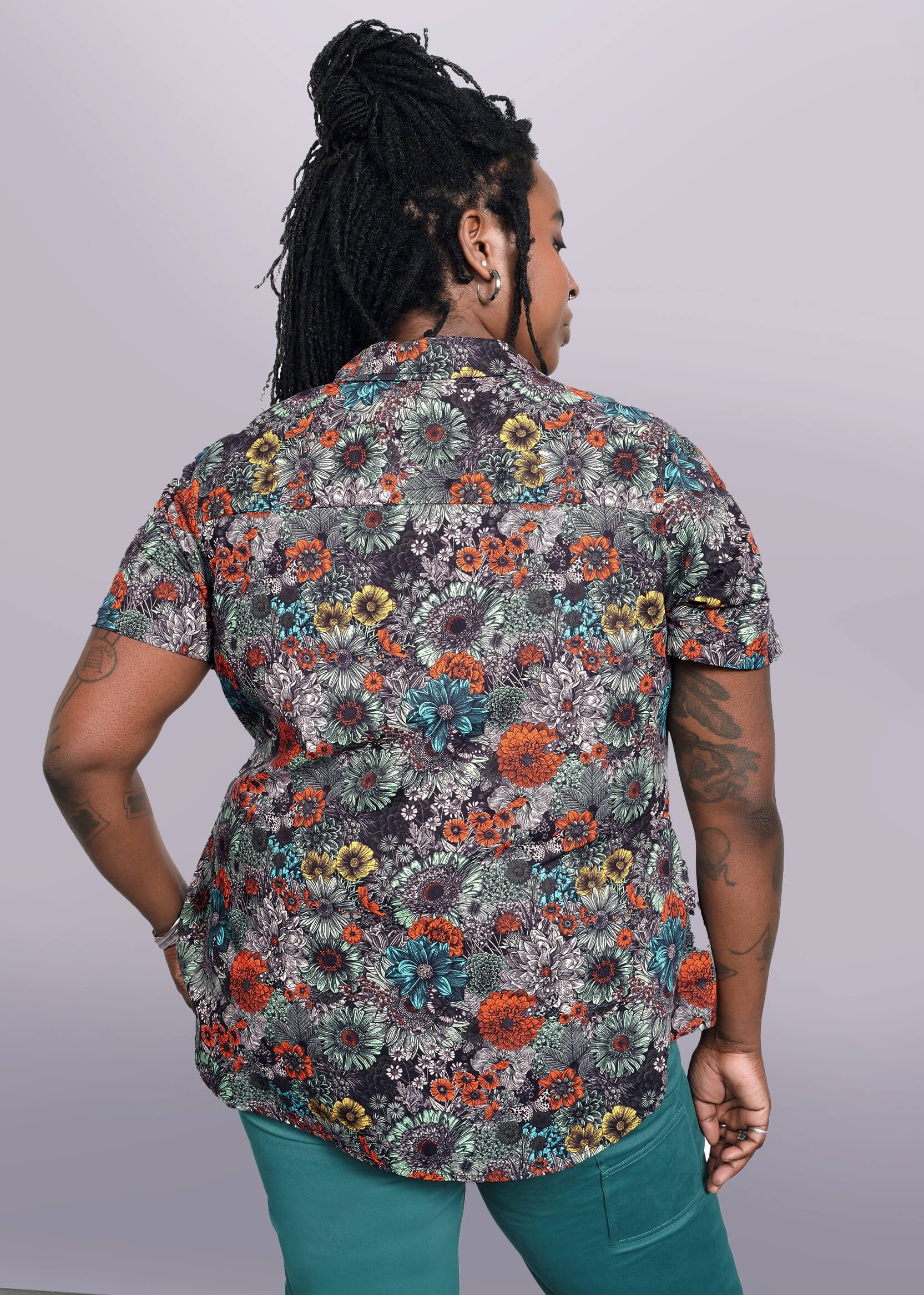 1X=A person with long braided hair and tattoos on their left arm is facing away, wearing The Essential Button Up, a short-sleeved floral cotton shirt with a classic collar and tailored fit, paired with teal pants. Their right hand rests on their hip against a plain white background.