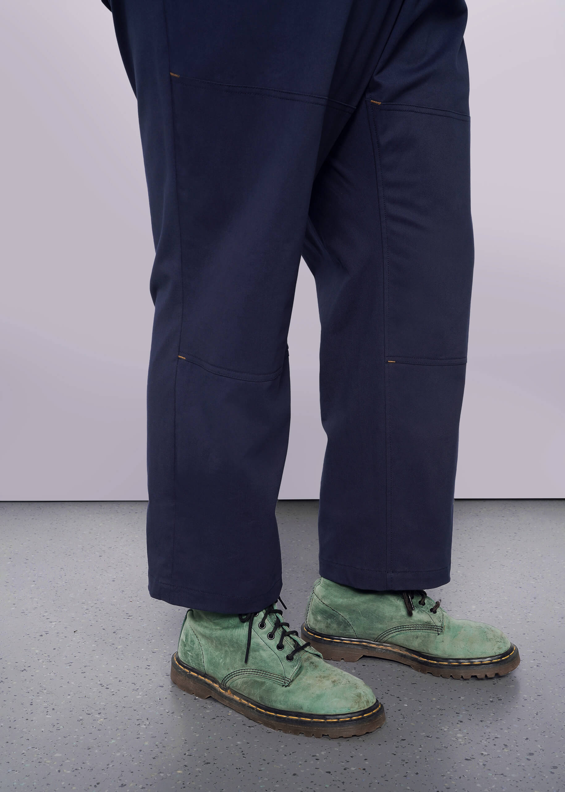 1X=A person wears The Essential Sleeveless High Waisted Coverall in dark blue with green lace-up suede boots, standing on a speckled gray floor. The white background accentuates the contrast between the cotton stretch coverall and the shoes.