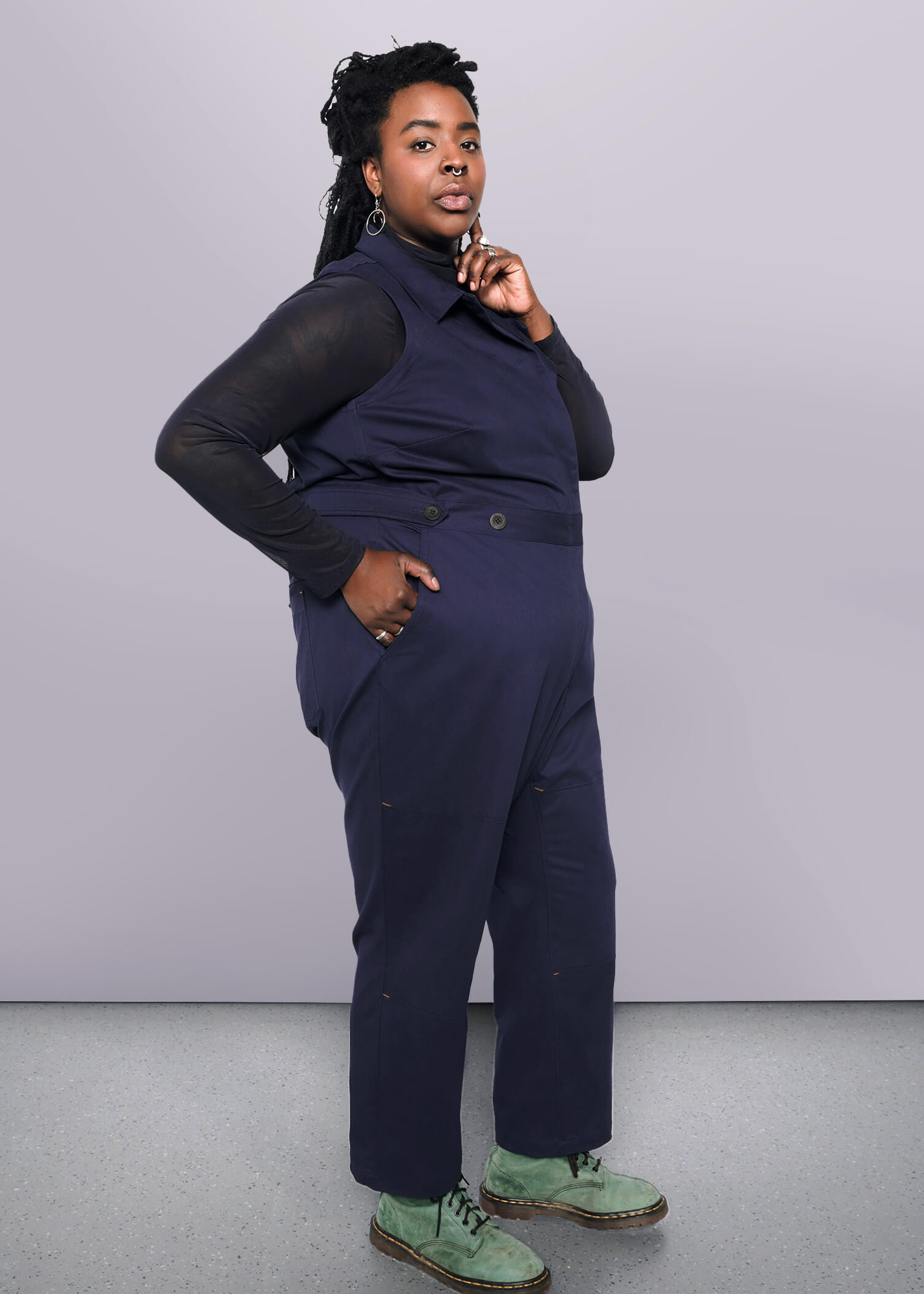 1X=A person confidently poses against a white background, wearing The Essential Sleeveless High Waisted Coverall in dark blue over a long-sleeve shirt and green shoes. With shoulder-length dark braided hair, they rest their chin on their hand.