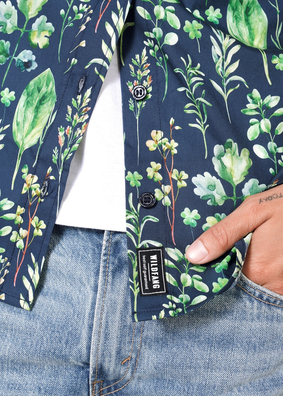 S=A person is wearing The Essential Button Up—a dark blue, short-sleeved 100% cotton shirt with a green leaf and botanical print. The shirt is partly buttoned over a white top, paired with light blue jeans. Their tattooed hands are visible. A clothing label reads WILDLIING.